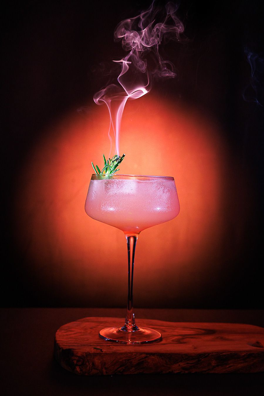 A smoky pink cocktail in a coupe glass garnished with rosemary, set against a dramatic orange-red backlit background.