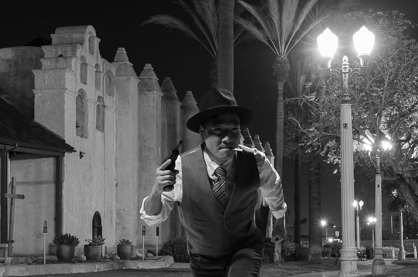 Man in hat and vest running at night near lit street lamp and white building in black and white photo.