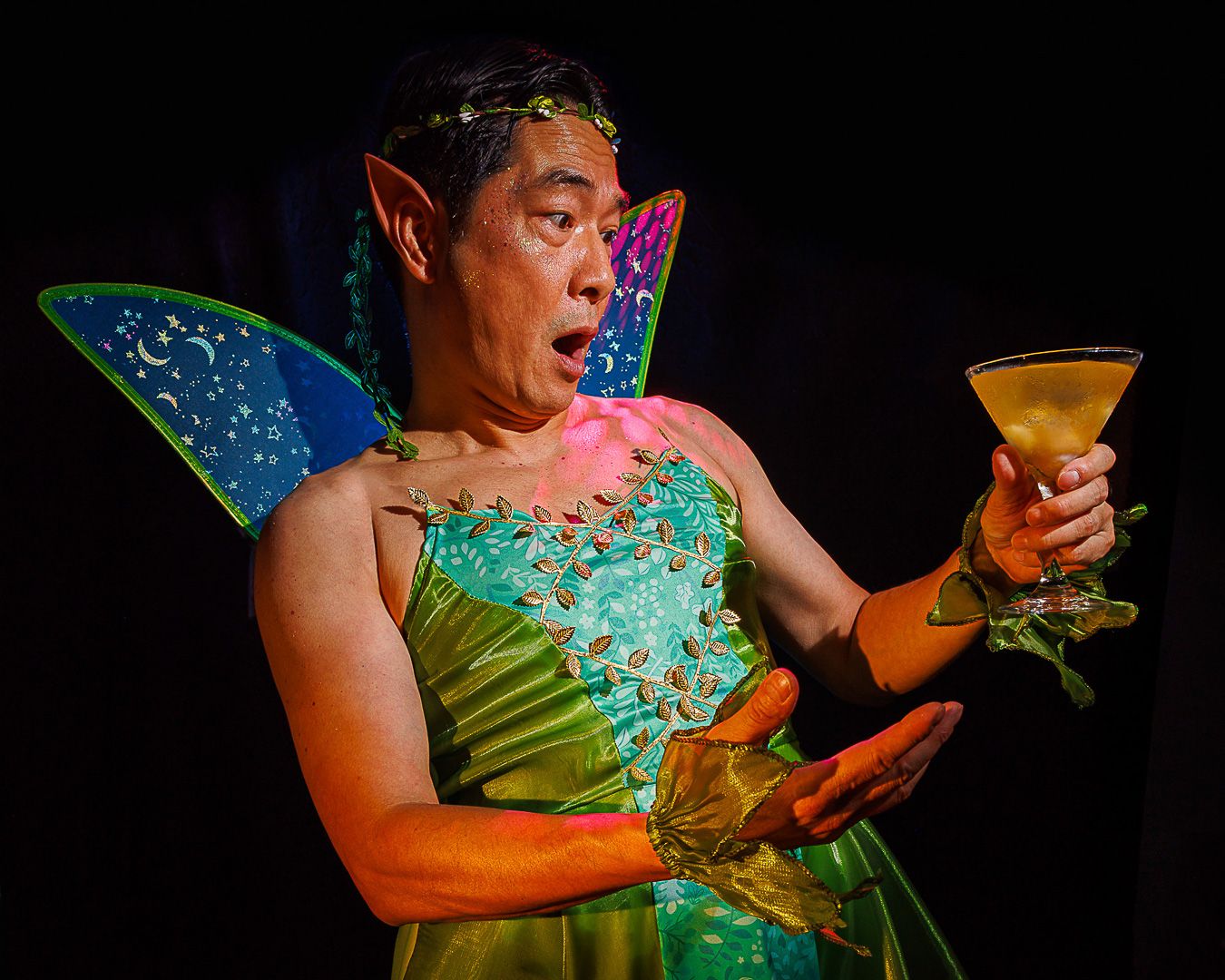 A performer in a green fairy costume with elf ears and wings holds a cocktail glass on stage.