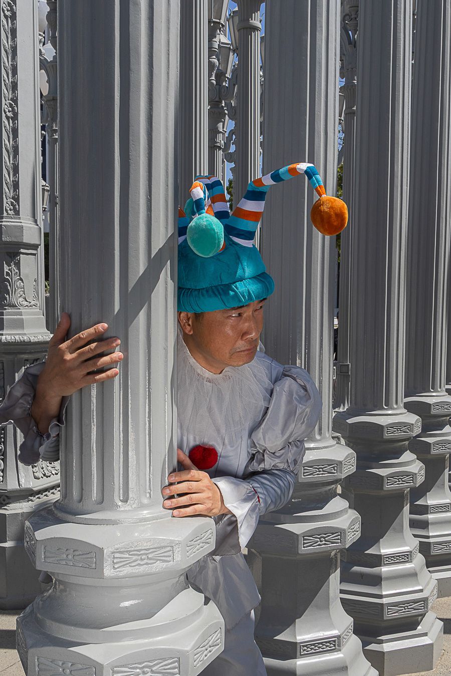 Person wearing colorful jester hat with pompoms poses between white ornate lamp posts in bright sunlight.