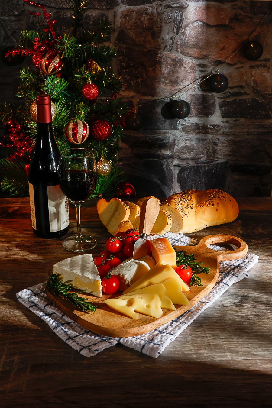 Festive Christmas cheese board with red wine, tomatoes, rosemary, and bread on rustic wooden table with decorated tree.