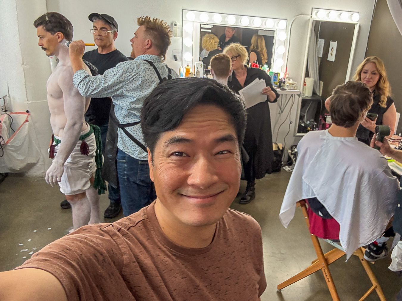 Man takes selfie in busy backstage dressing room with performers and makeup artists preparing in the background.