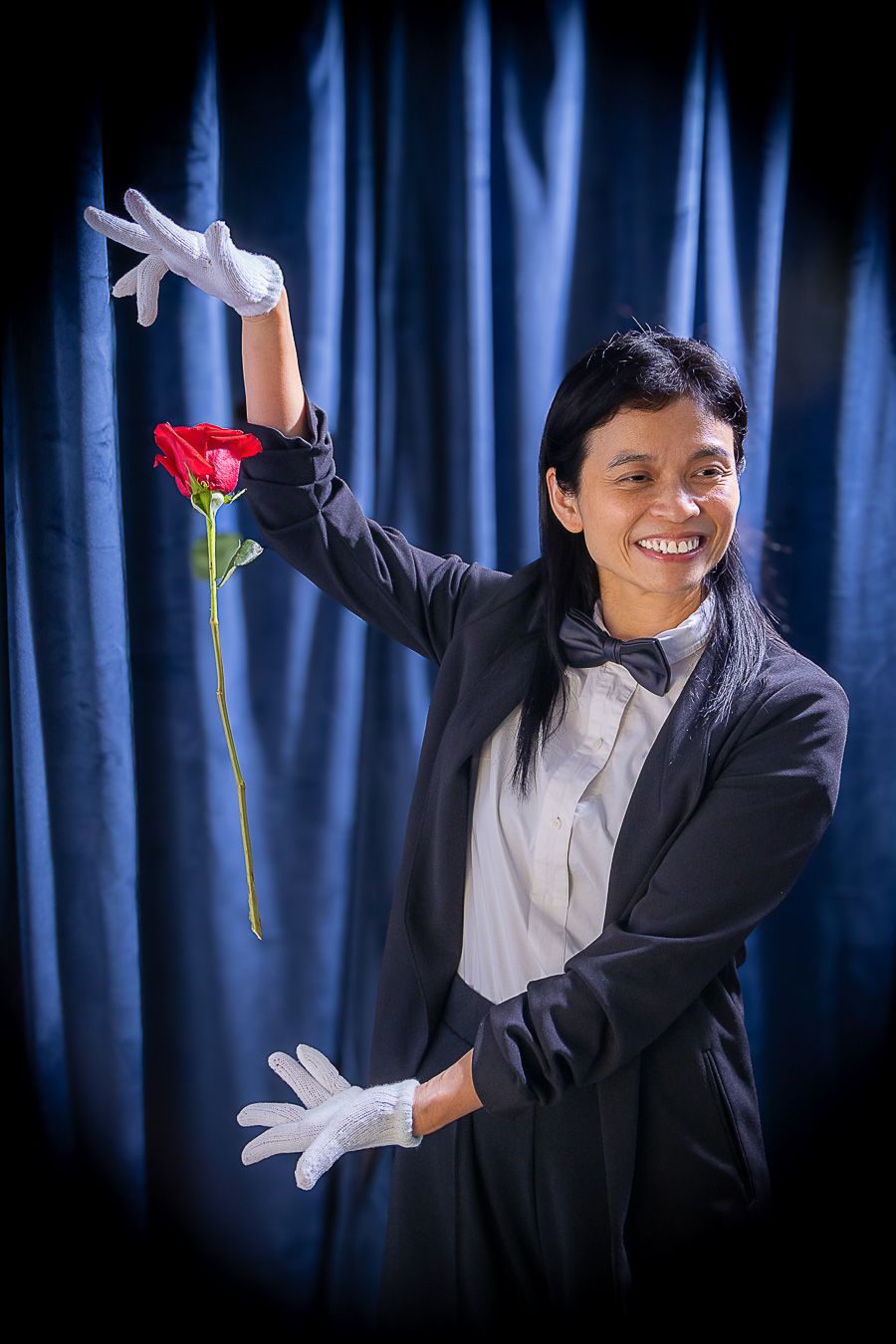 Smiling magician in tuxedo and white gloves presenting a red rose on stage with blue curtain backdrop.
