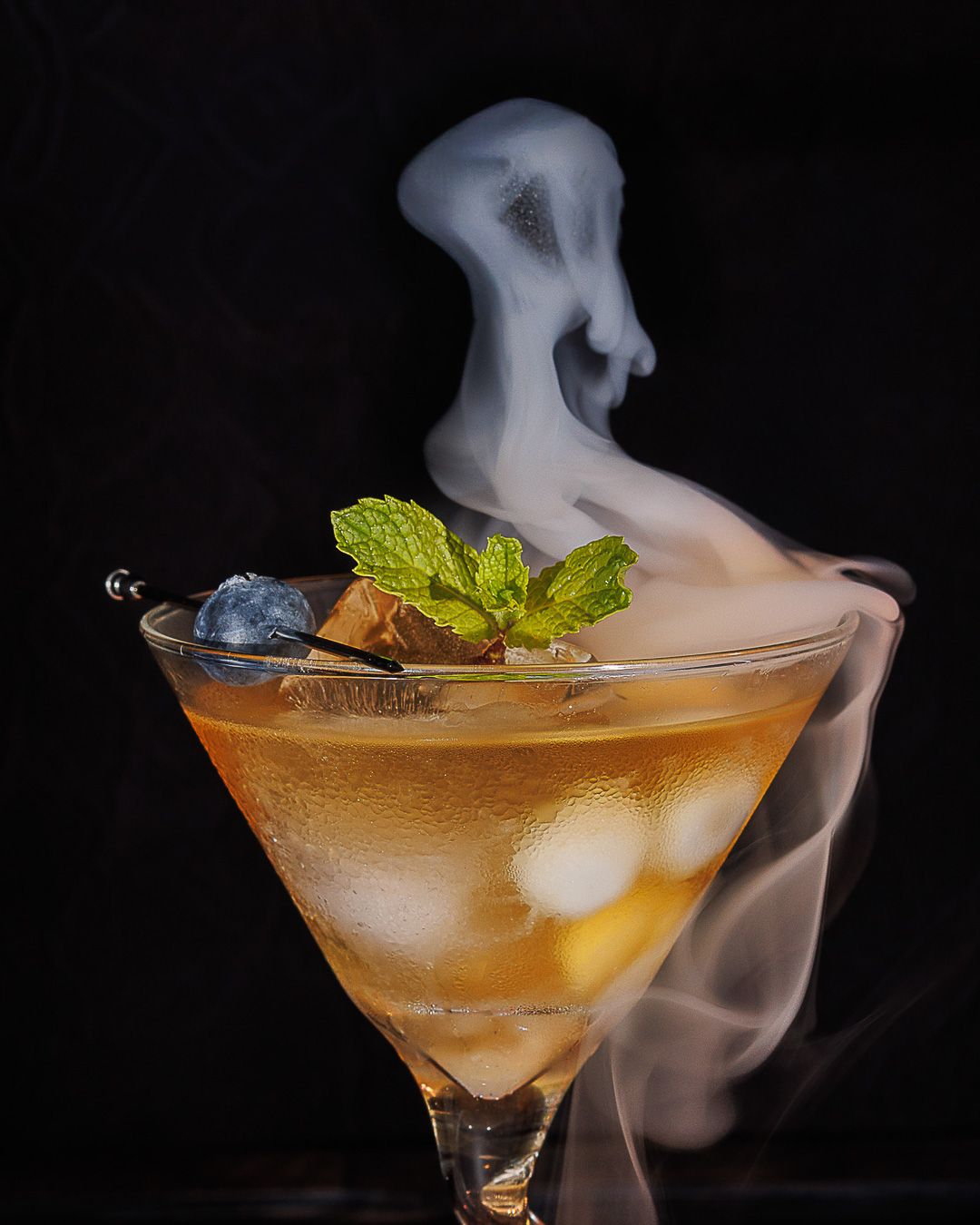 Smoky Halloween cocktail in martini glass with mint garnish, ice, and ghost-shaped smoke rising against black background.