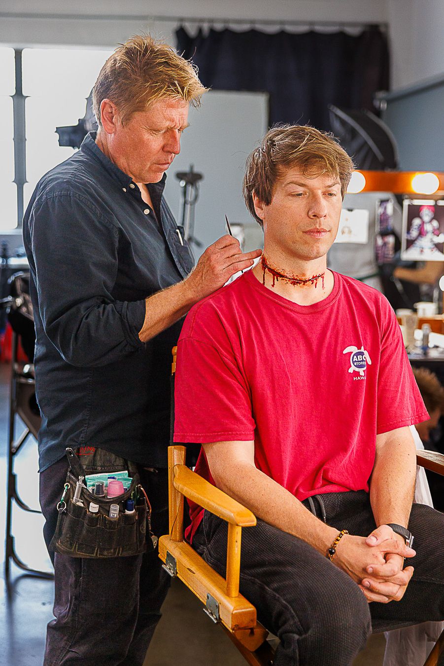 Makeup artist applying special effects wound makeup to a man in a red shirt seated in a director's chair.