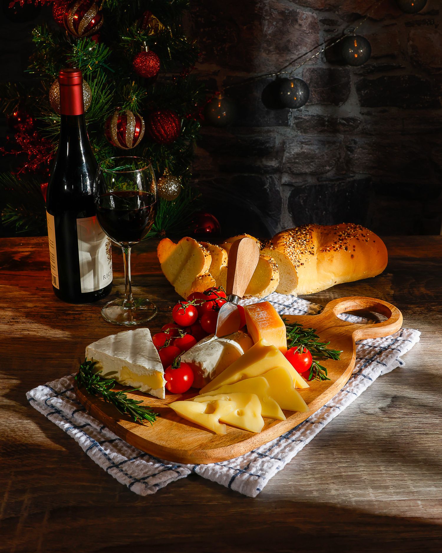 Holiday cheese board with wine, tomatoes, rosemary, and bread near Christmas tree.