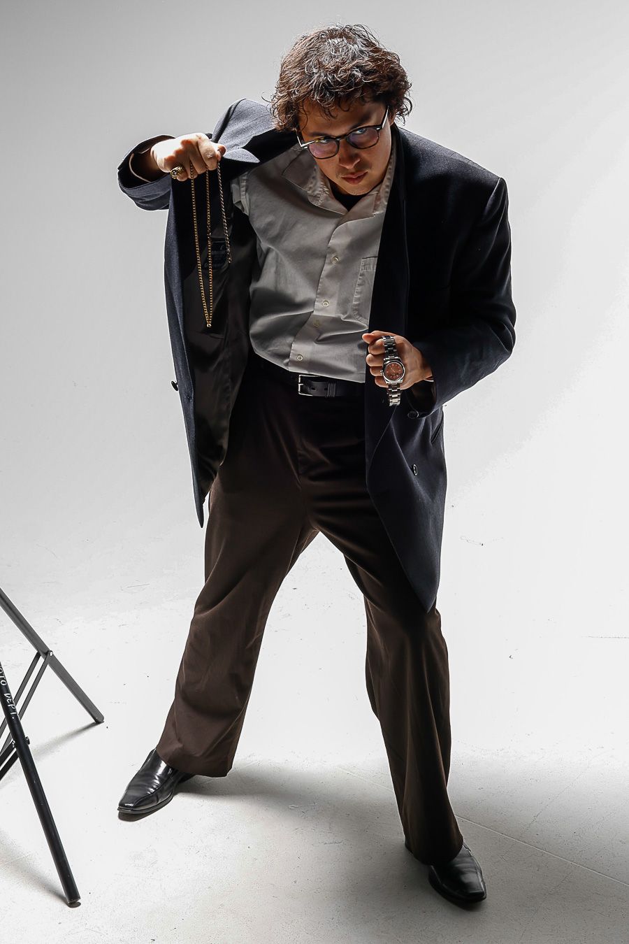 A man in a dark suit and glasses holds a pocket watch while reaching into his jacket on a white background.