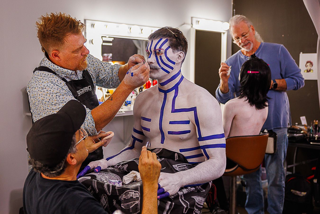 Artists apply intricate blue and white body paint designs on a performer backstage under bright makeup lights.