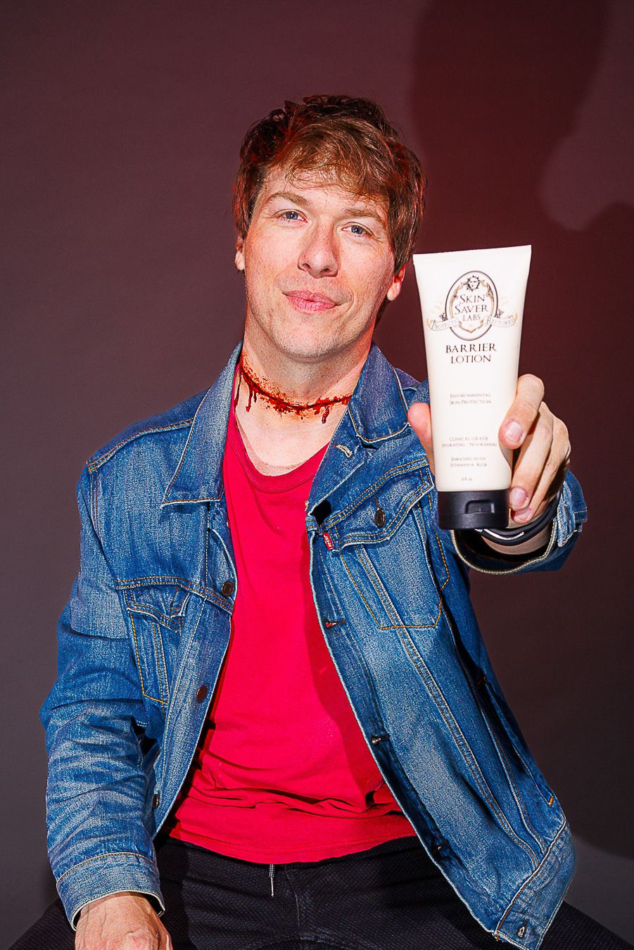 Man in denim jacket and red shirt holds a barrier lotion tube, with theatrical fake blood on his neck.