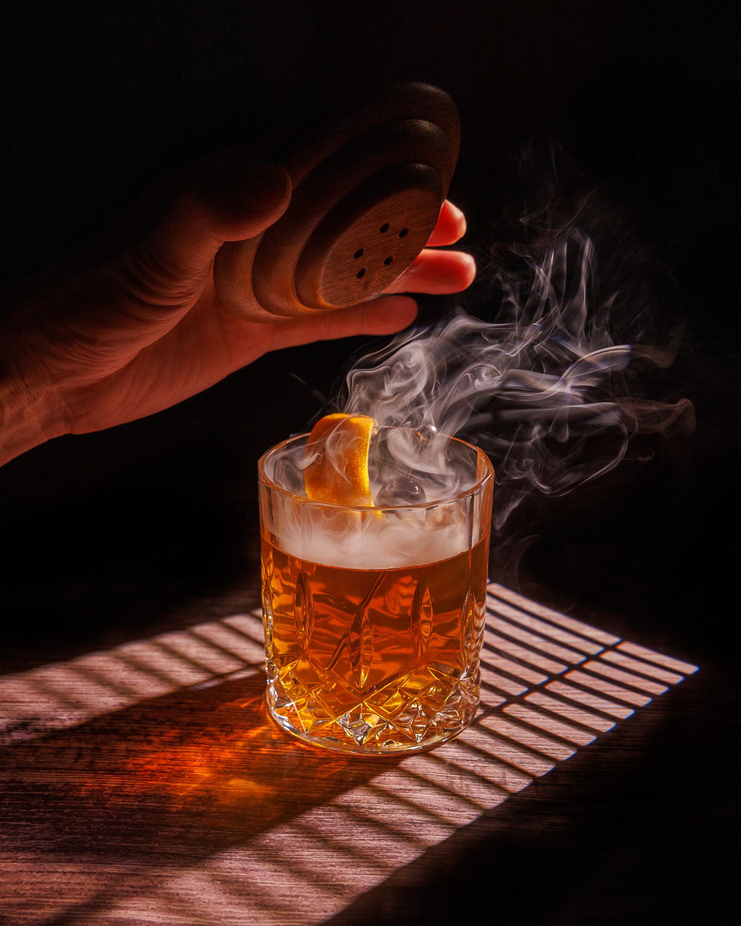 A hand holding a smoking gun over a smoky whiskey cocktail with a lemon twist in a crystal rocks glass.