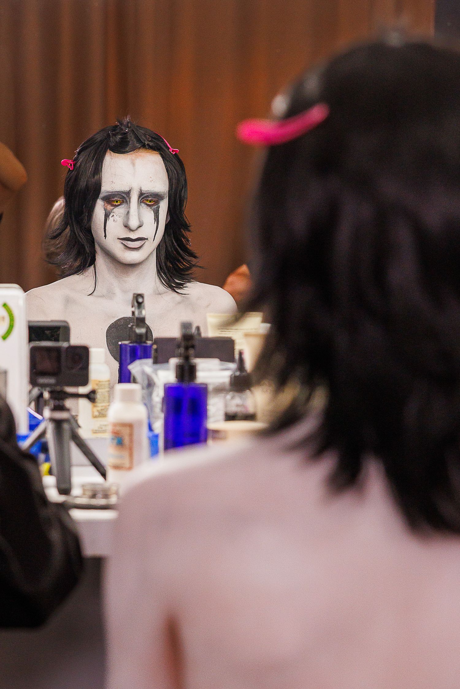 Performer with dramatic white face paint and dark eye makeup sits before a mirror surrounded by makeup products backstage.
