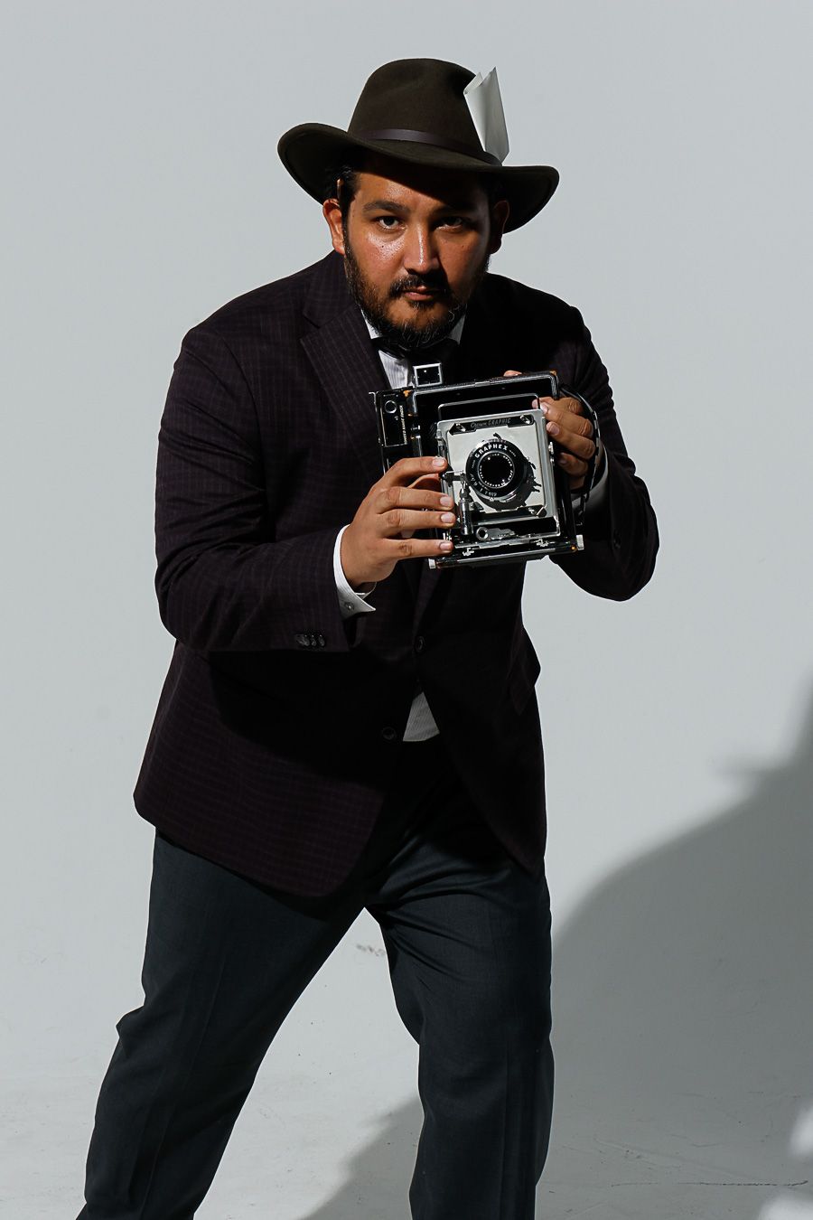 Man in dark suit and fedora hat holding vintage large format camera, posing as retro press photographer on gray background.