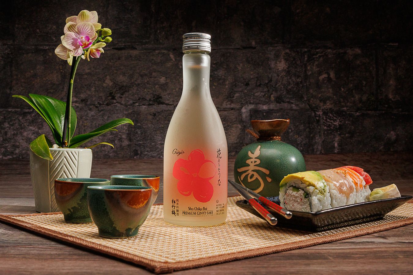 Japanese sake bottle with sushi roll, green ceramic cups, vase, and pink orchid on bamboo mat.