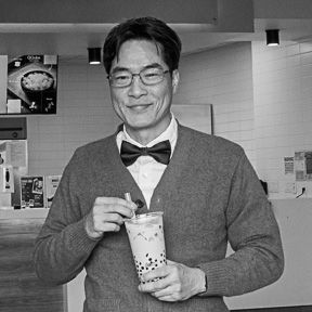 Man in bow tie smiling while holding bubble tea drink in a cafe setting.