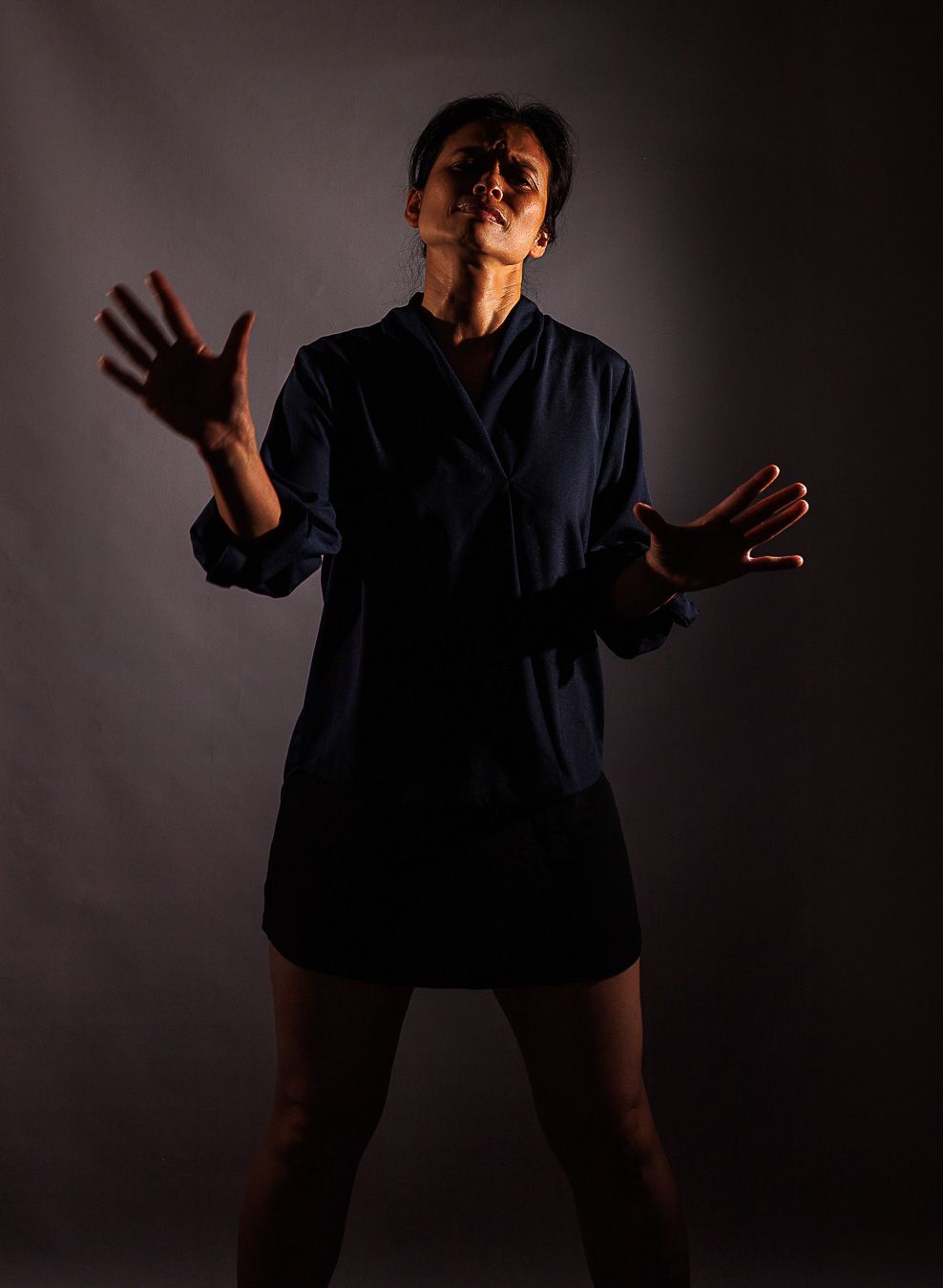 A woman in a dark blue dress shirt poses dramatically with arms outstretched against a dark studio background.