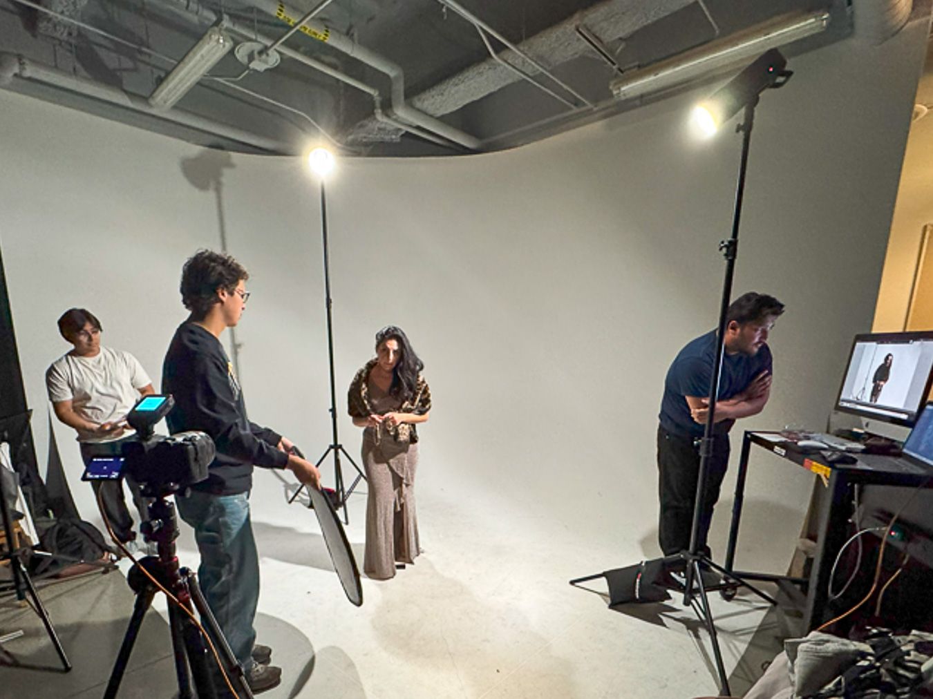 Photography studio shoot with crew members, lighting equipment, and a female model posing against a white cyclorama backdrop.