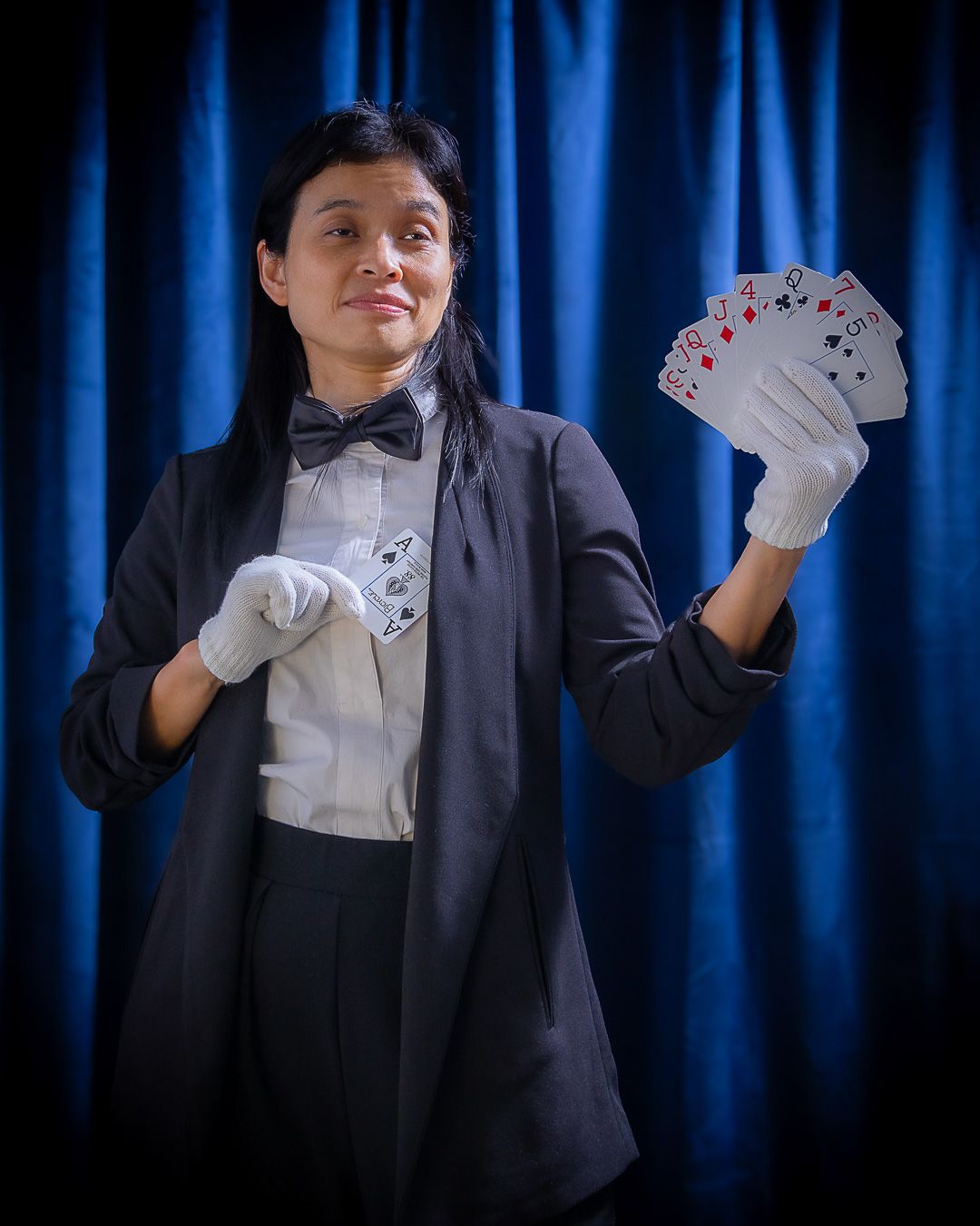Female magician in tuxedo holding fanned playing cards wearing white gloves against blue curtain backdrop.