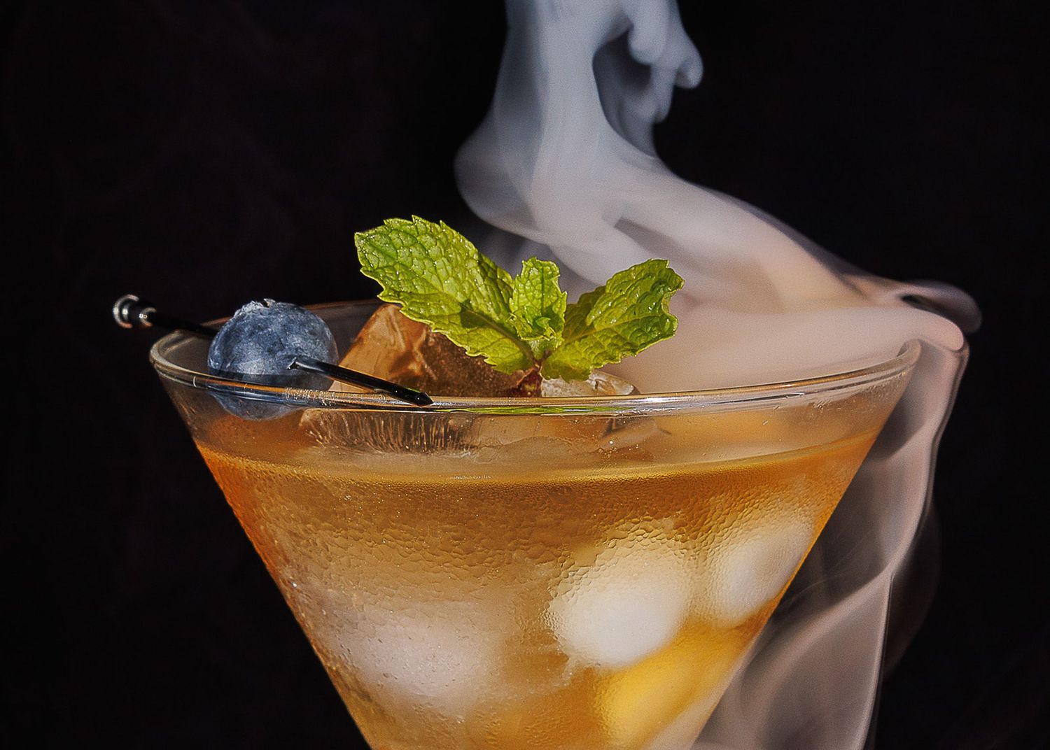 A smoky cocktail in a martini glass with ice, fresh mint garnish, and a stirrer on a dark background.