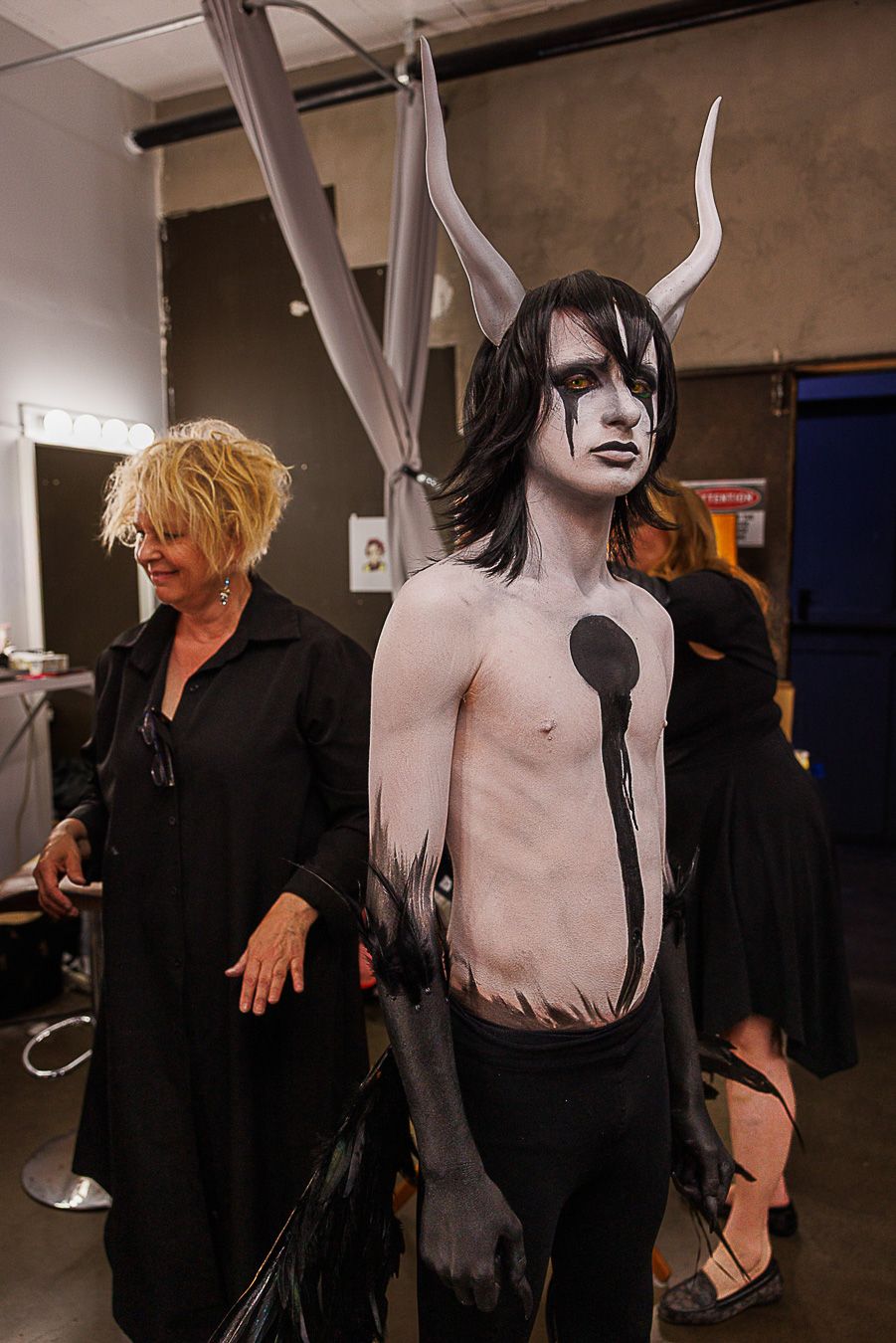 Performer in dramatic demon costume with white body paint, black horns, and dark makeup backstage with makeup artist.