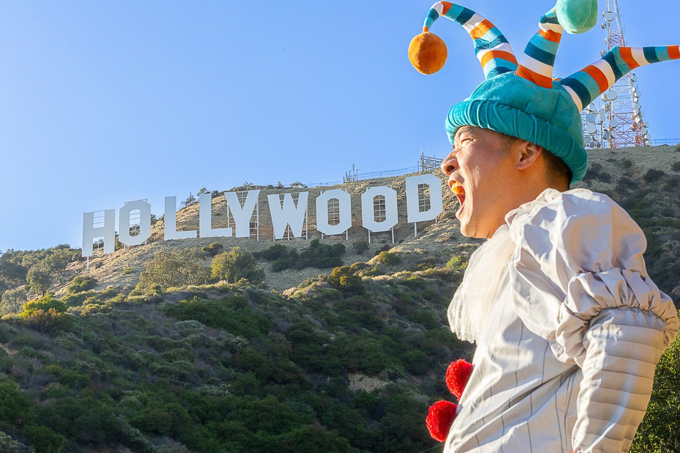 Person in colorful jester hat laughing in front of the iconic Hollywood Sign on a sunny day in Los Angeles.