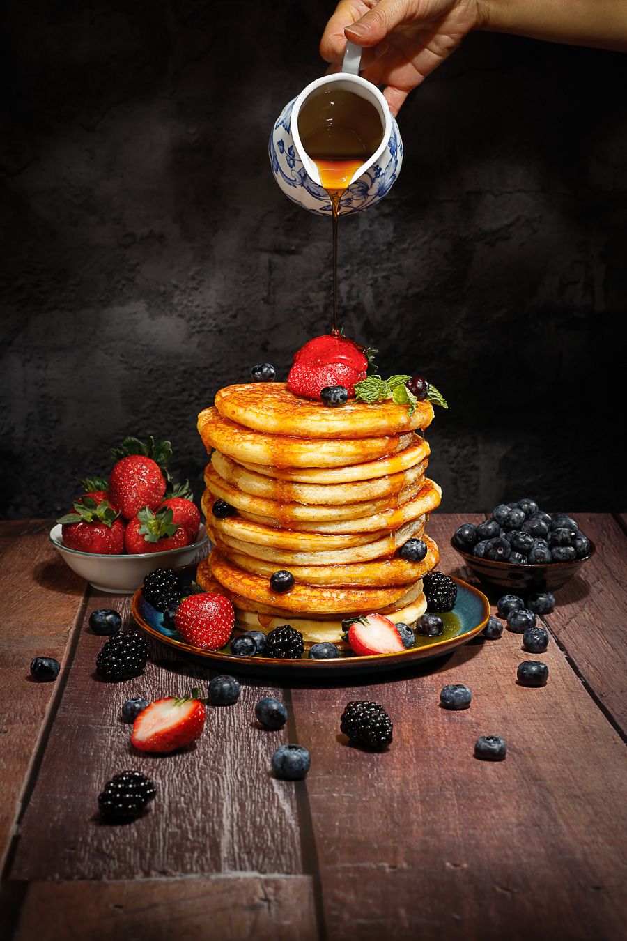 Stack of fluffy pancakes topped with fresh strawberries, blueberries, blackberries, and maple syrup being poured from a pitcher.
