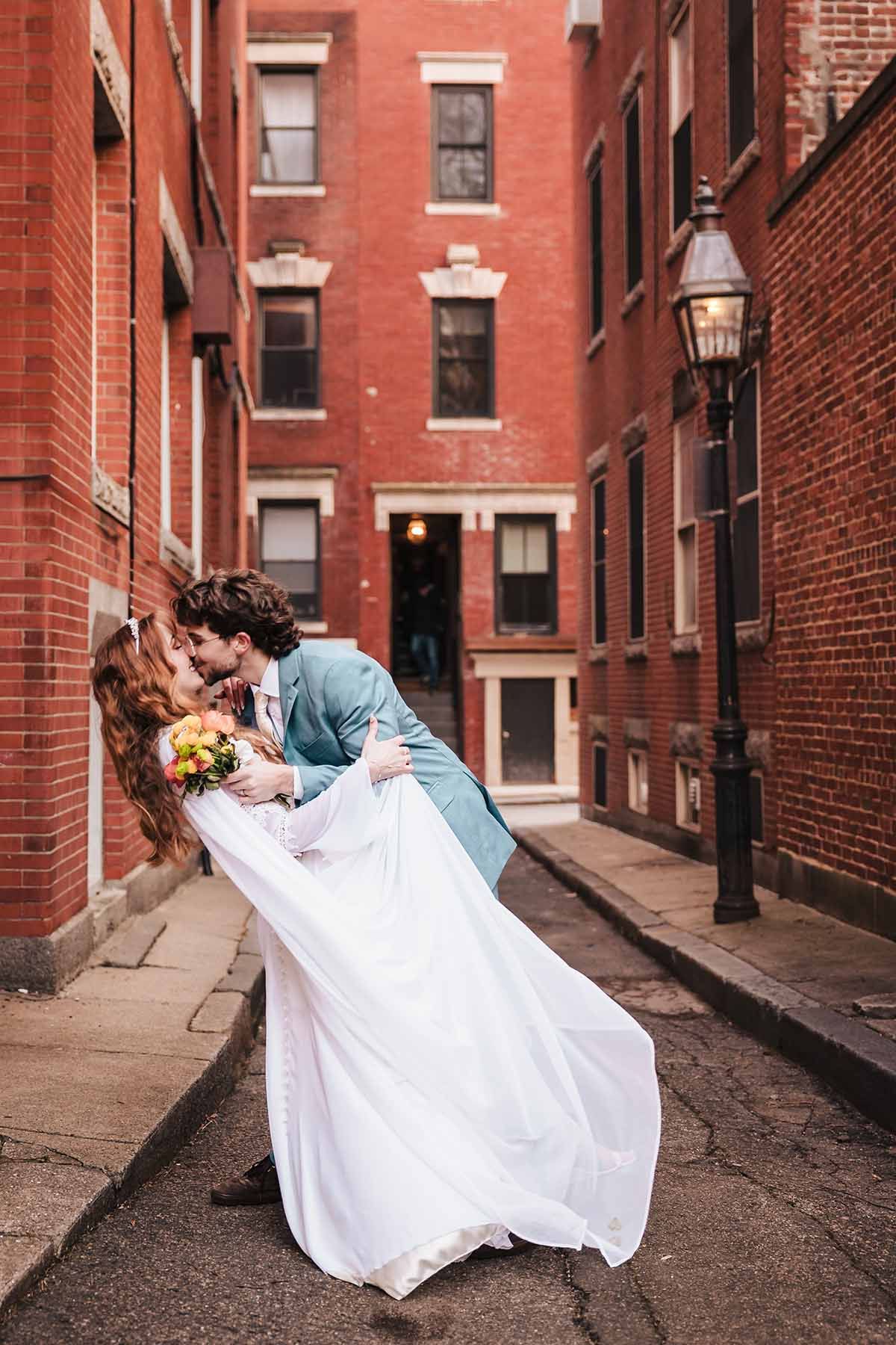 Groom carries bride through a narrow brick alleyway in the North End area of Boston, her white dress flowing behind them during their elope