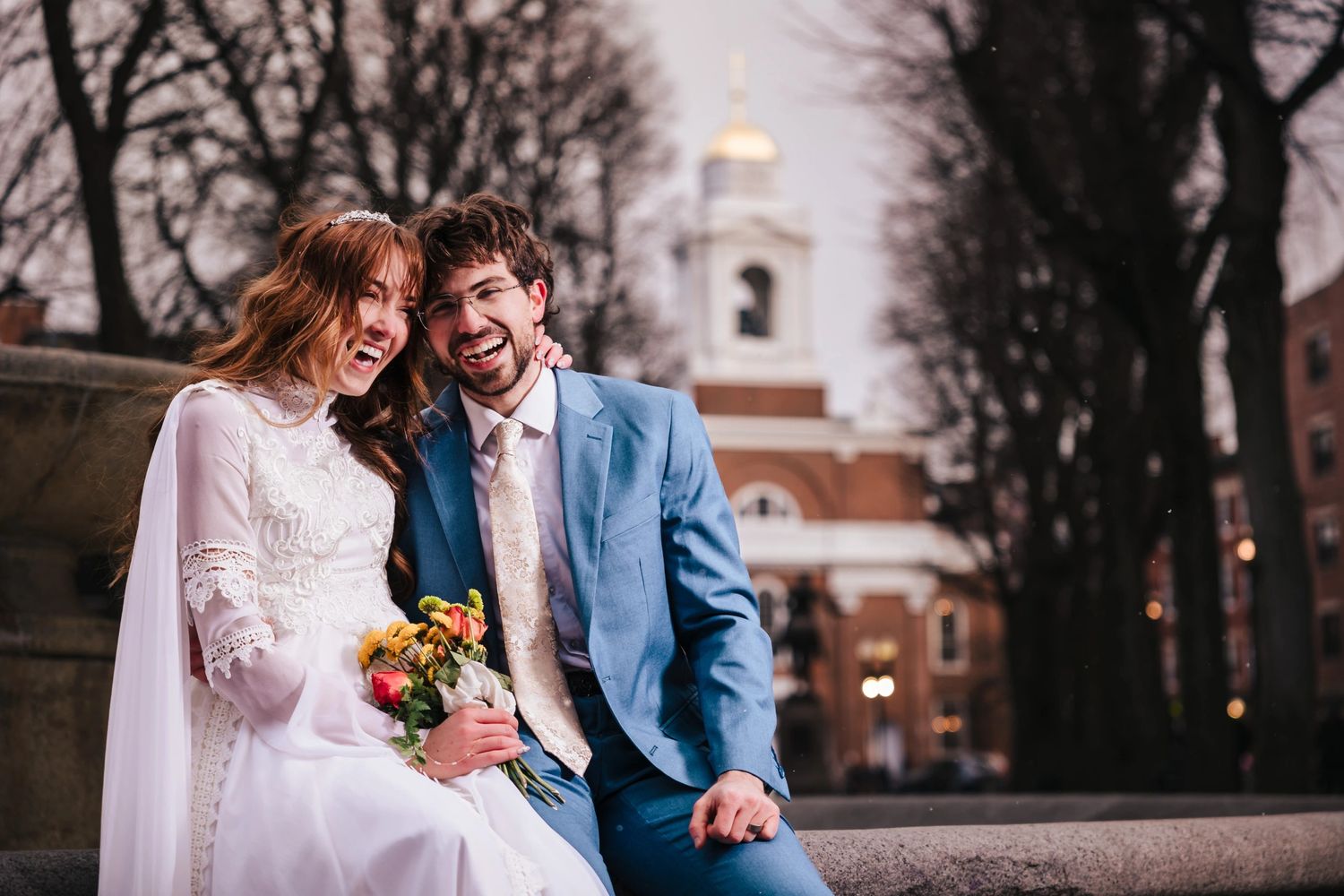 Bride and groom sit together on a ledge in their wedding attire, smiling at Paul Revere Mall, during their Boston Elopement Session.
