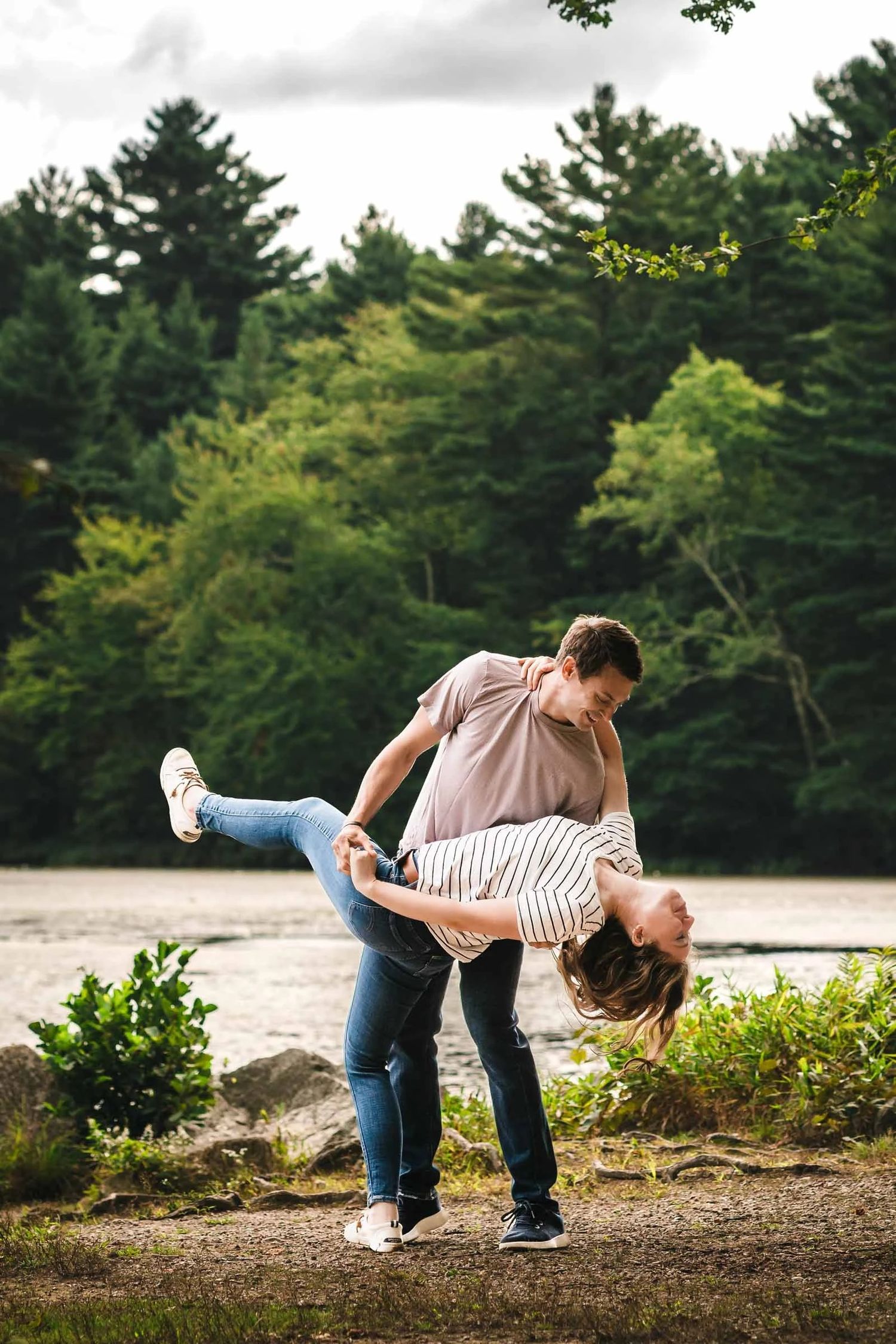 Man playfully lifting woman off the ground in a spinning embrace by a lakeside. Engagement at Borderland State Park, North Easton, MA