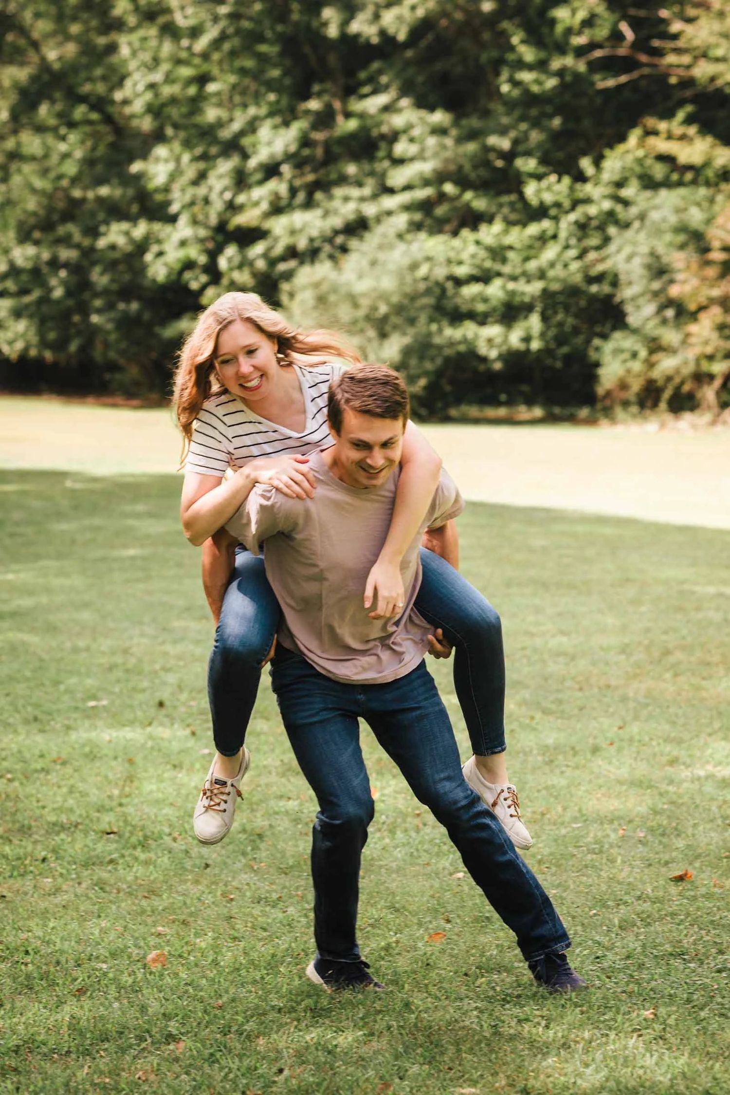 Man giving woman a piggyback ride across a grassy lawn, both smiling and playful. Engagement at Borderland State Park, North Easton, MA