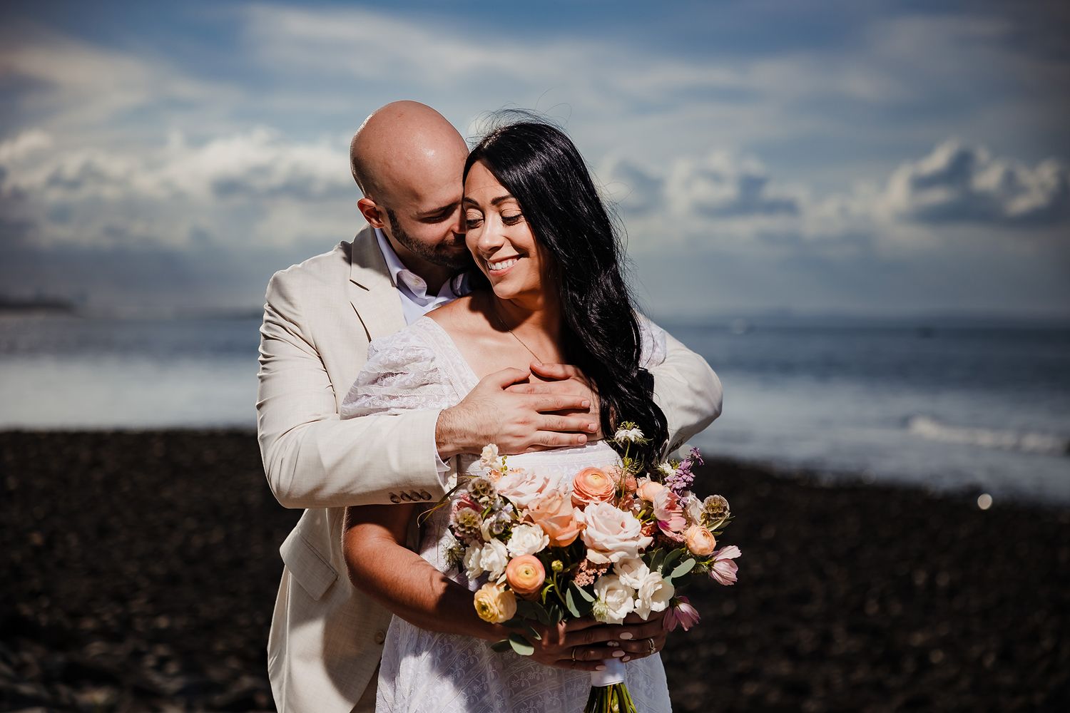 The bride and groom share a close, affectionate, embrace, on the beach in the Town of Hull MA with the Atlantic behind t