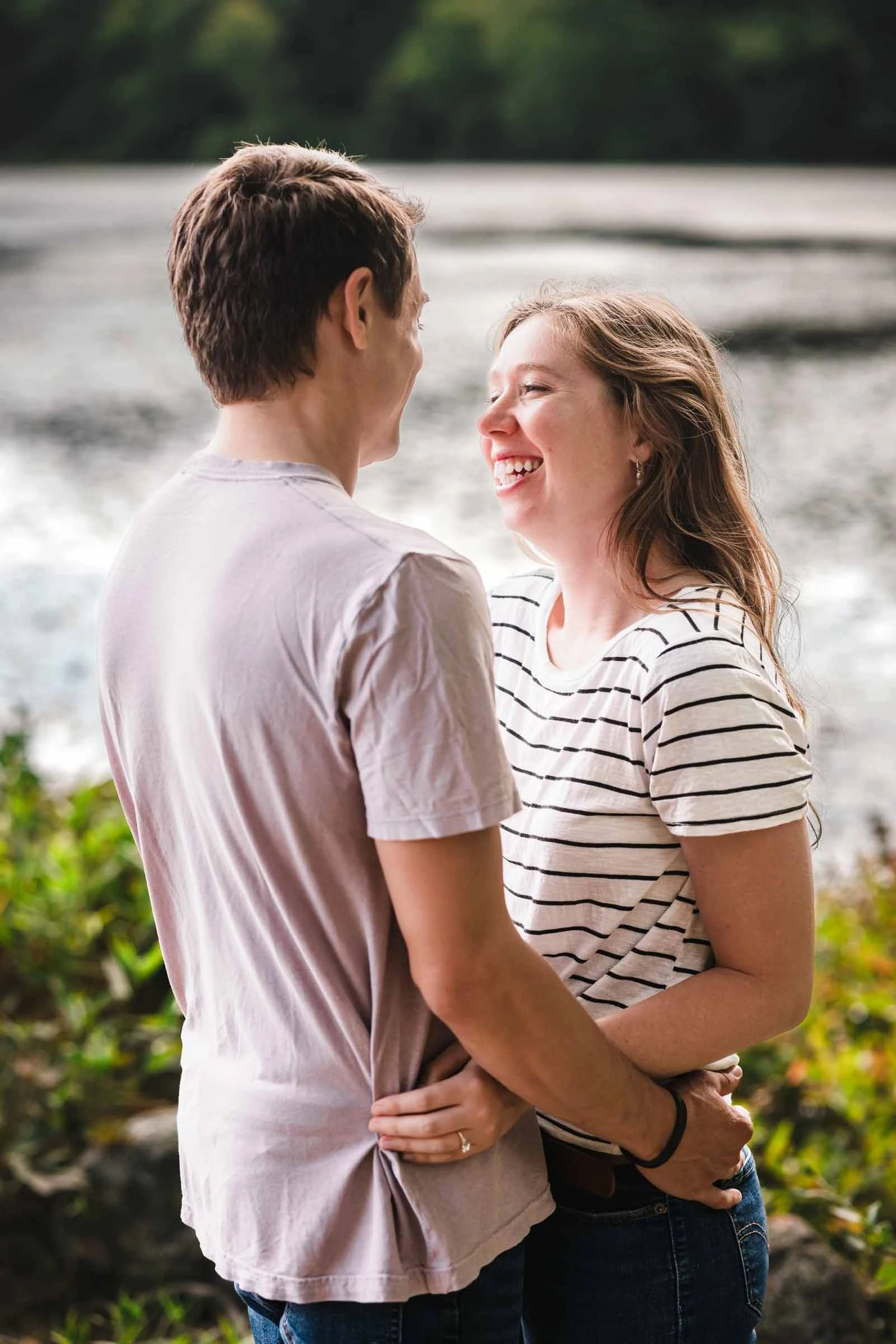 Couple embracing and smiling at each other by the water's edge during golden hour. Engagement at Borderland State Park, North Easton, MA