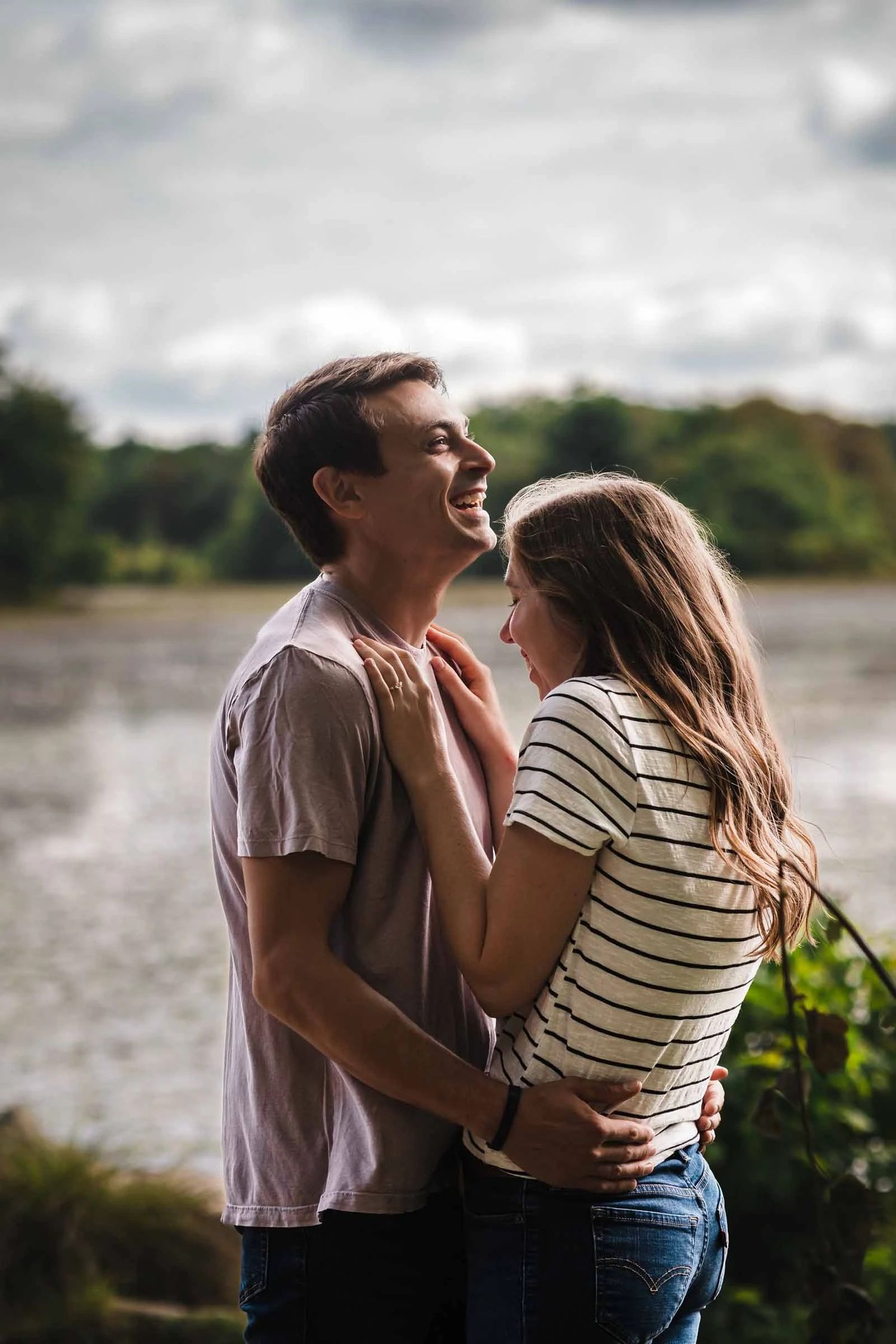 Couple embracing and gazing toward the horizon by a riverbank. Engagement at Borderland State Park, North Easton, MA