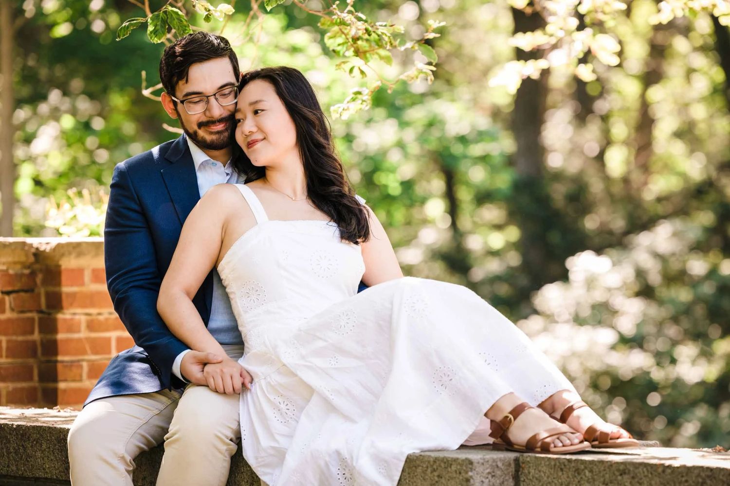 Couple sitting on stone wall outdoors, woman in white dress, man in blue blazer, surrounded by lush green trees.