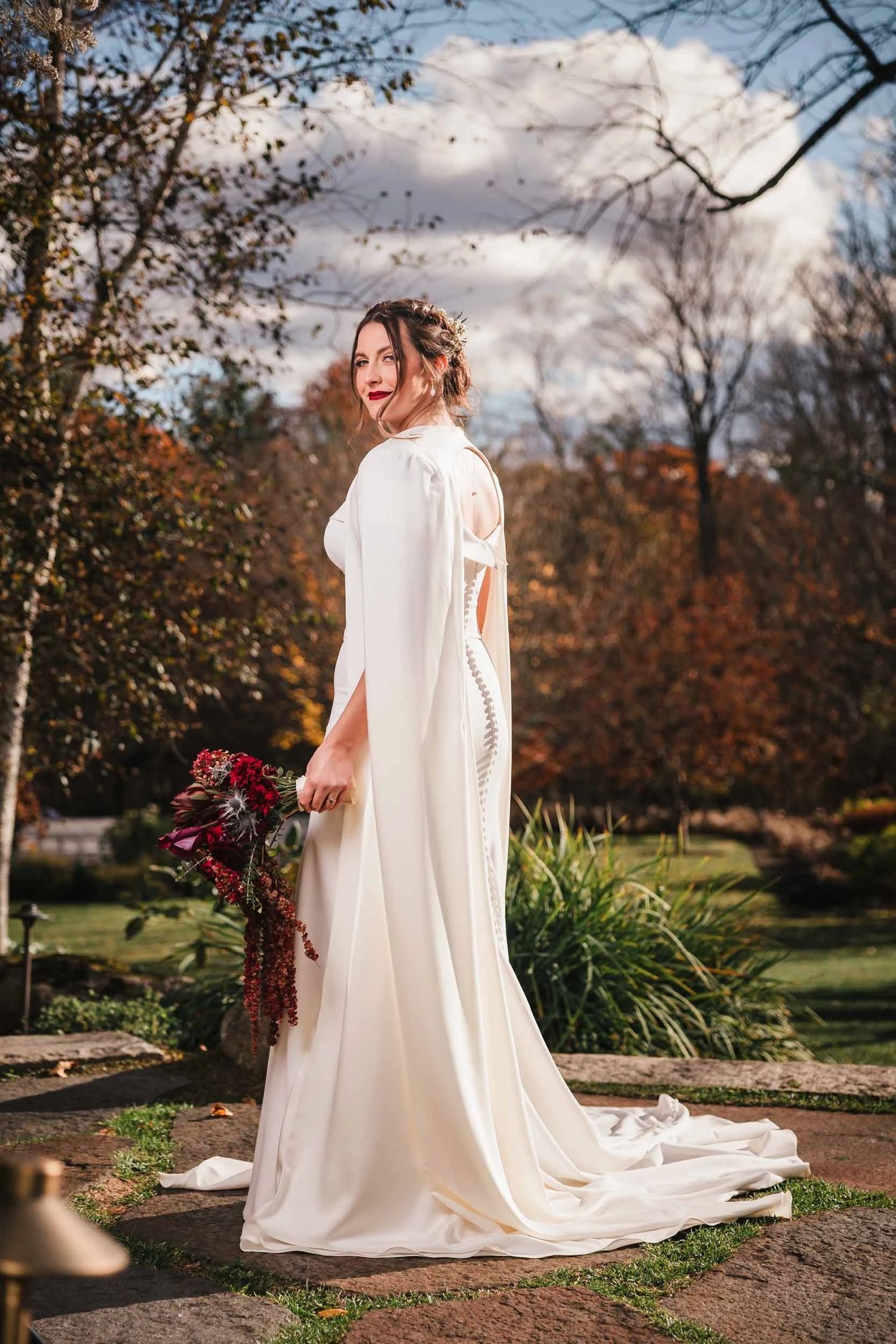 Bride standing alone in a garden, looking over her shoulder with a gentle smile, holding a deep red bouquet. Boston Wedding Photographer.