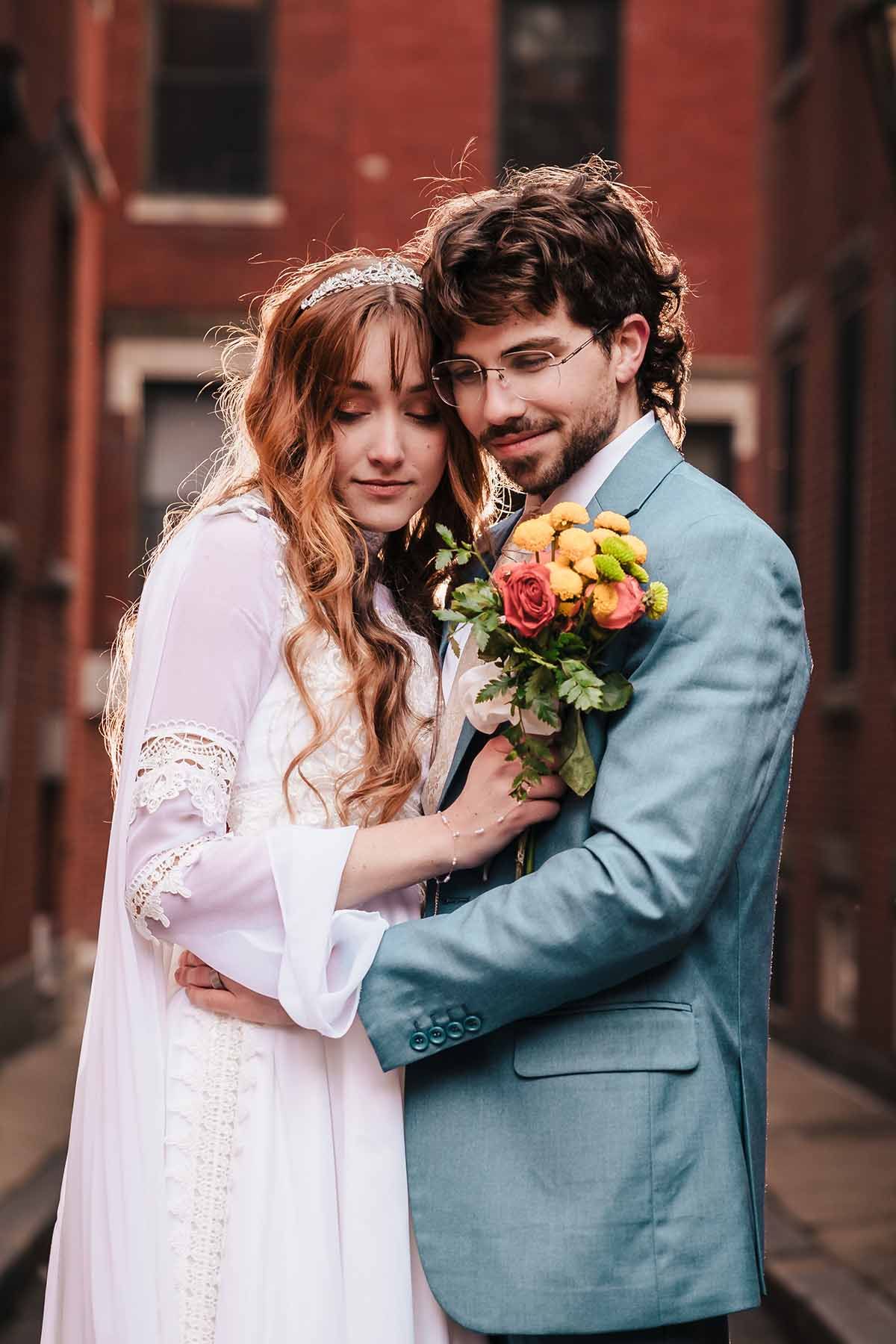 Bride in white lace gown embraces groom in blue suit holding colorful bouquet, posing outdoors in the North End of Boston