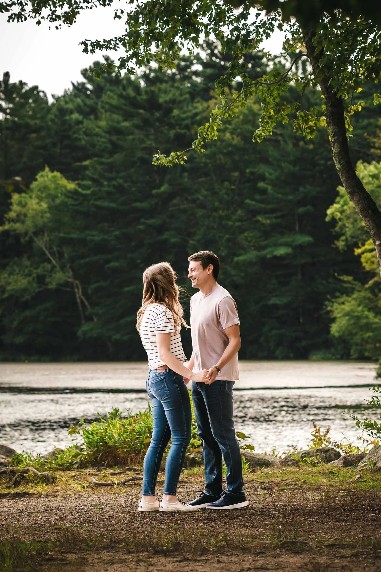 Young couple standing together by a river, facing each other in conversation.  Engagement at Borderland State Park, North Easton, MA