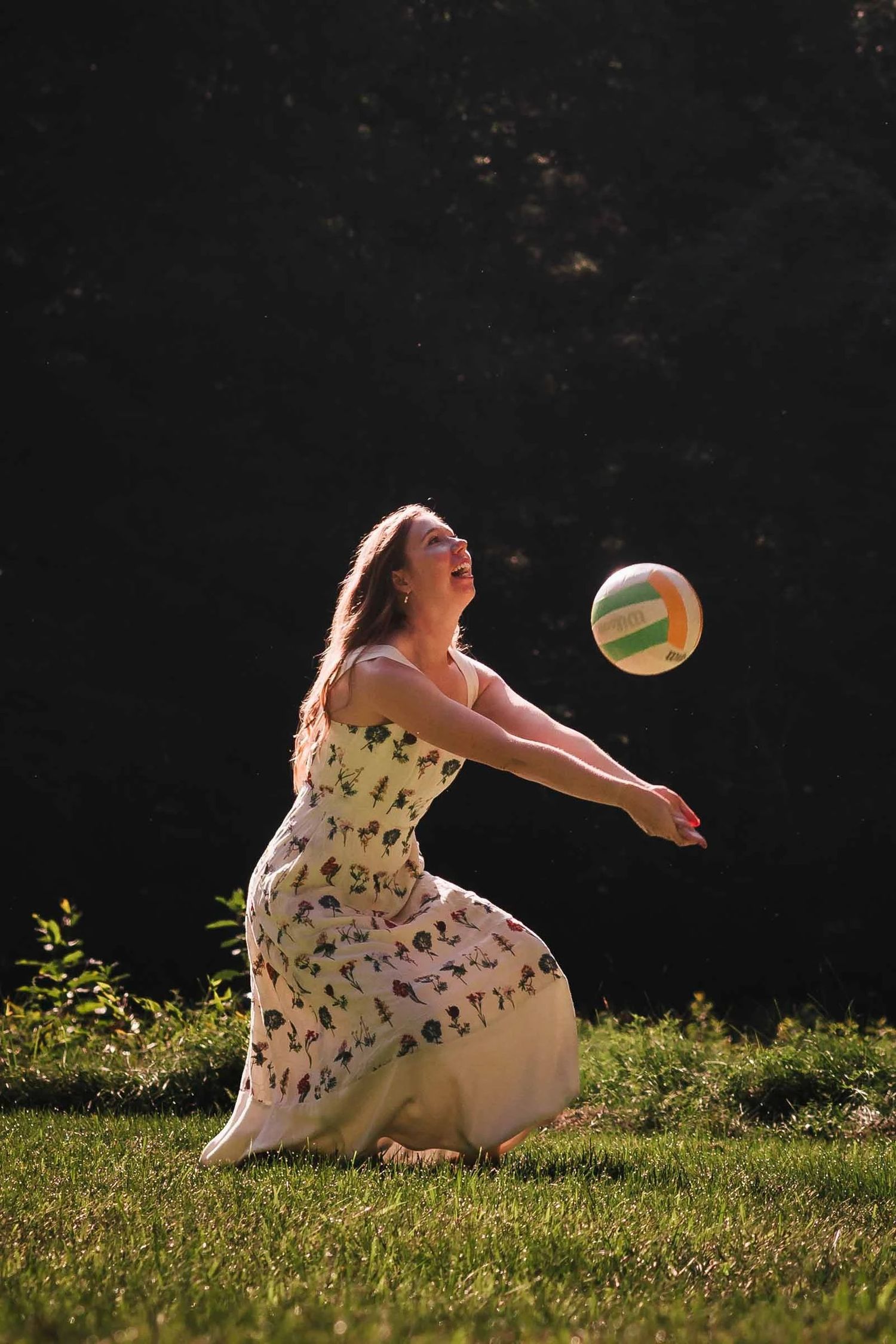 Woman kneeling on grass, passing a volleyball to herself in a backlit outdoor moment. Engagement at Borderland State Park, North Easton