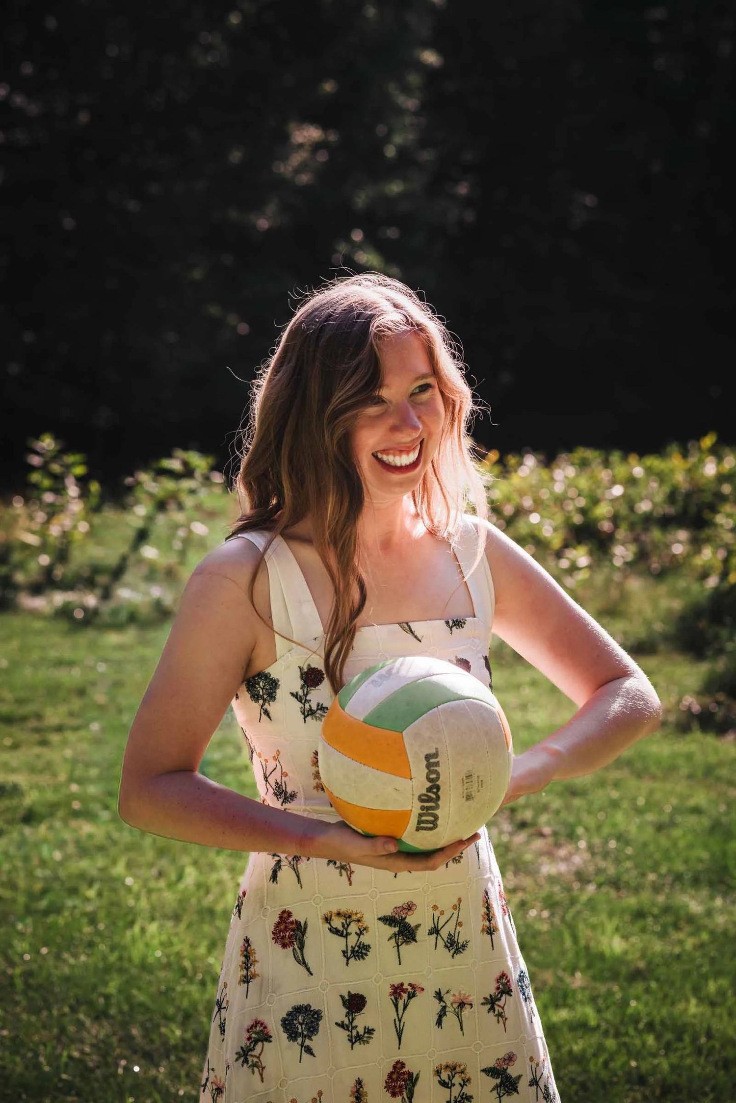 Woman holding a volleyball, smiling at the camera in a sunlit garden. Engagement at Borderland State Park, North Easton, MA