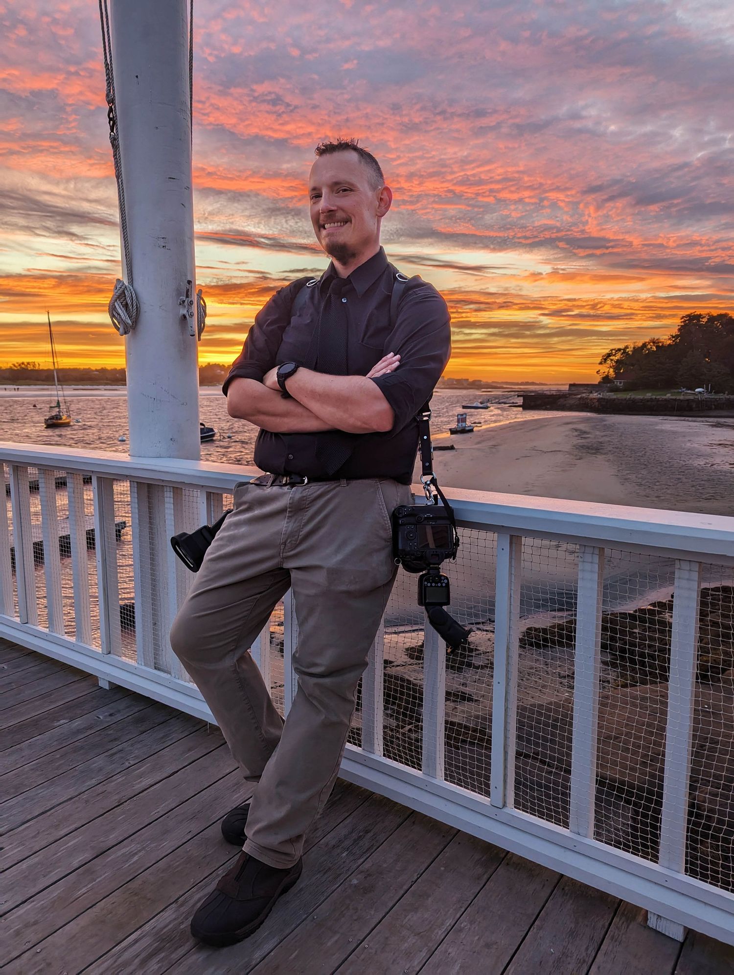 Boston wedding photographer Gregory Hitchcock standing on a pier at sunset, smiling with arms crossed and cameras at his sides.