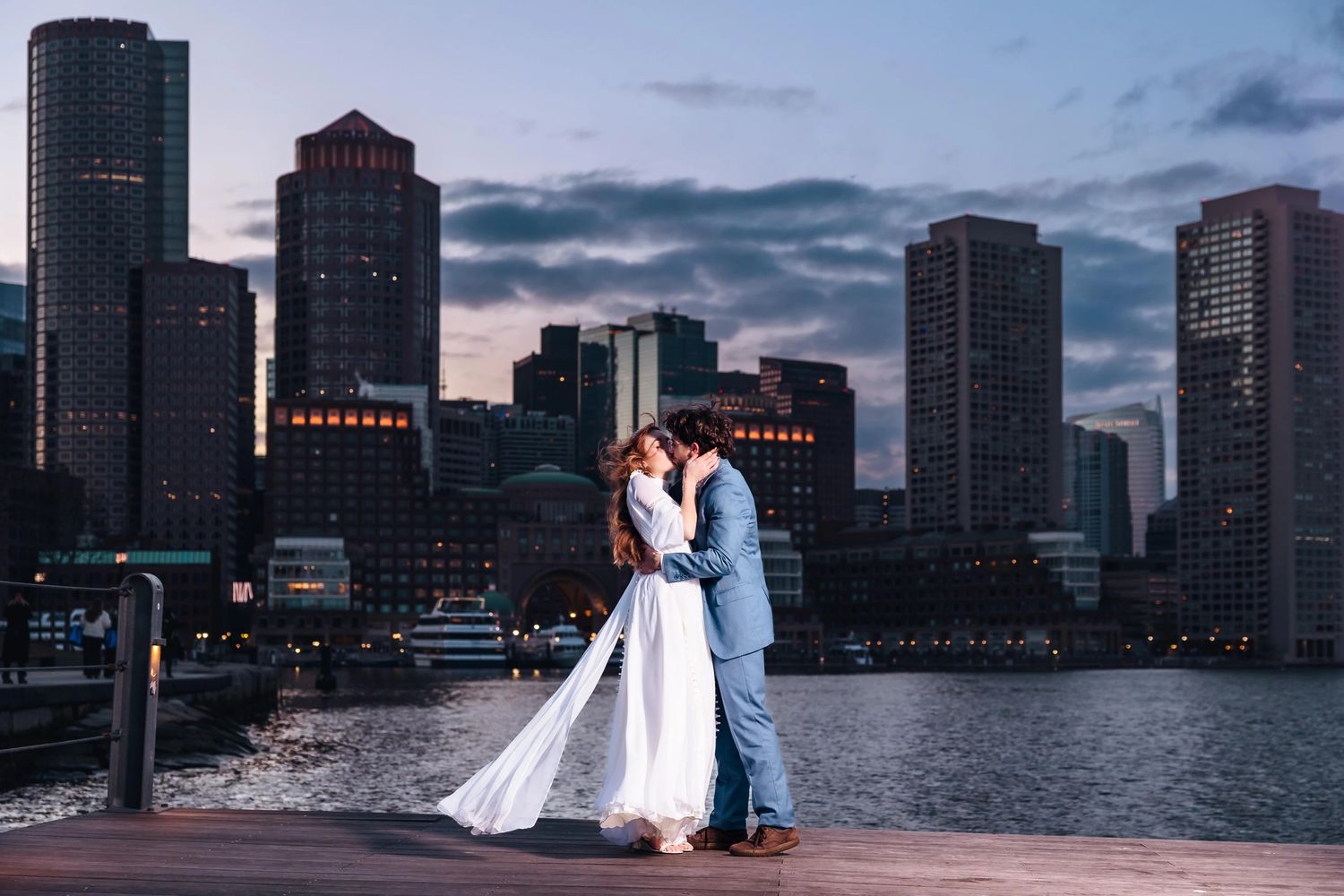 Bride and groom kiss on the marina at Fan Pier Park, at dusk during their Boston elopement, city lights glowing behind them.