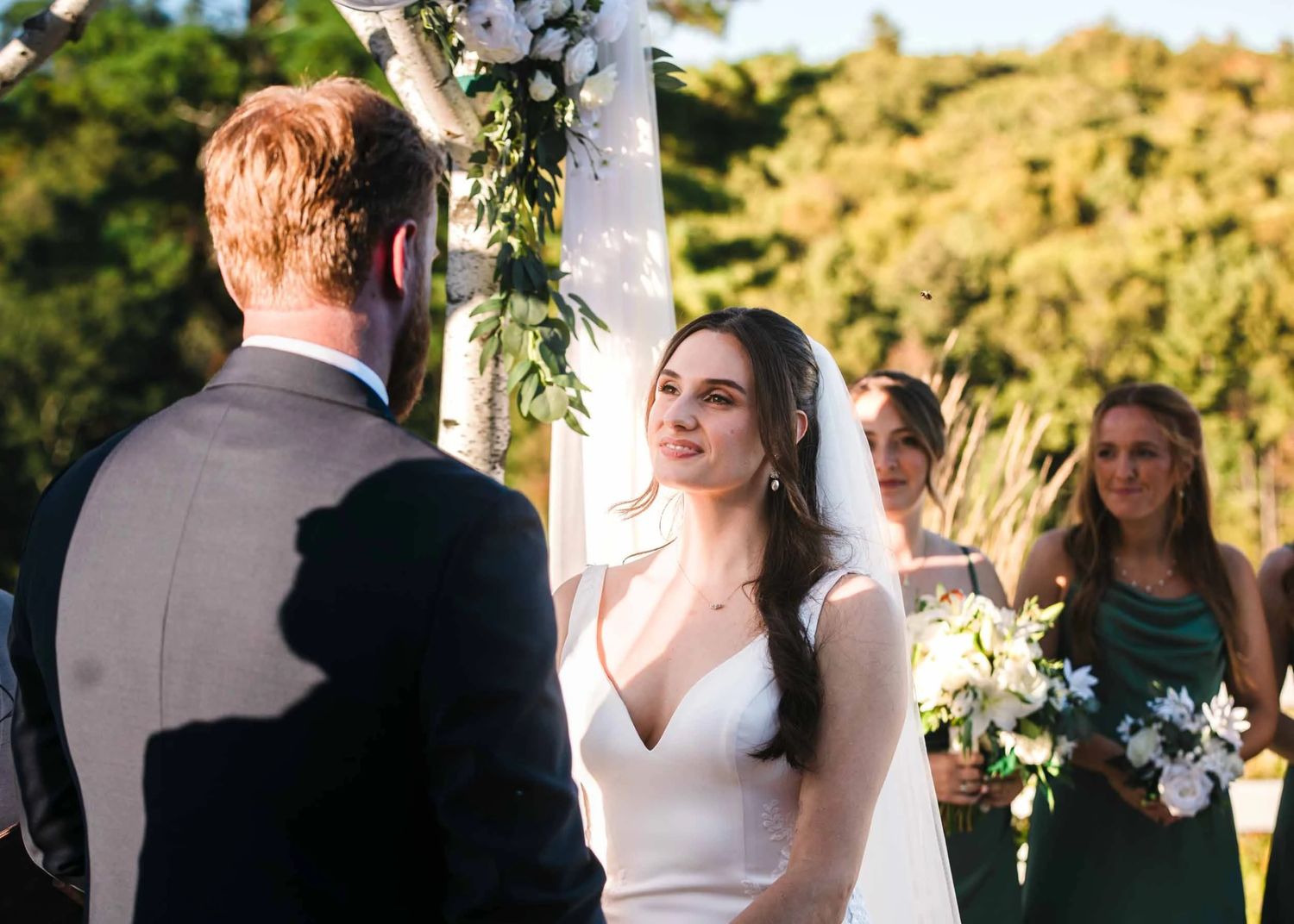 A beauitful bride gazes into her soon to be husband's eyes during a outdoor summer wedding ceremony with her bridesmaids behind her.