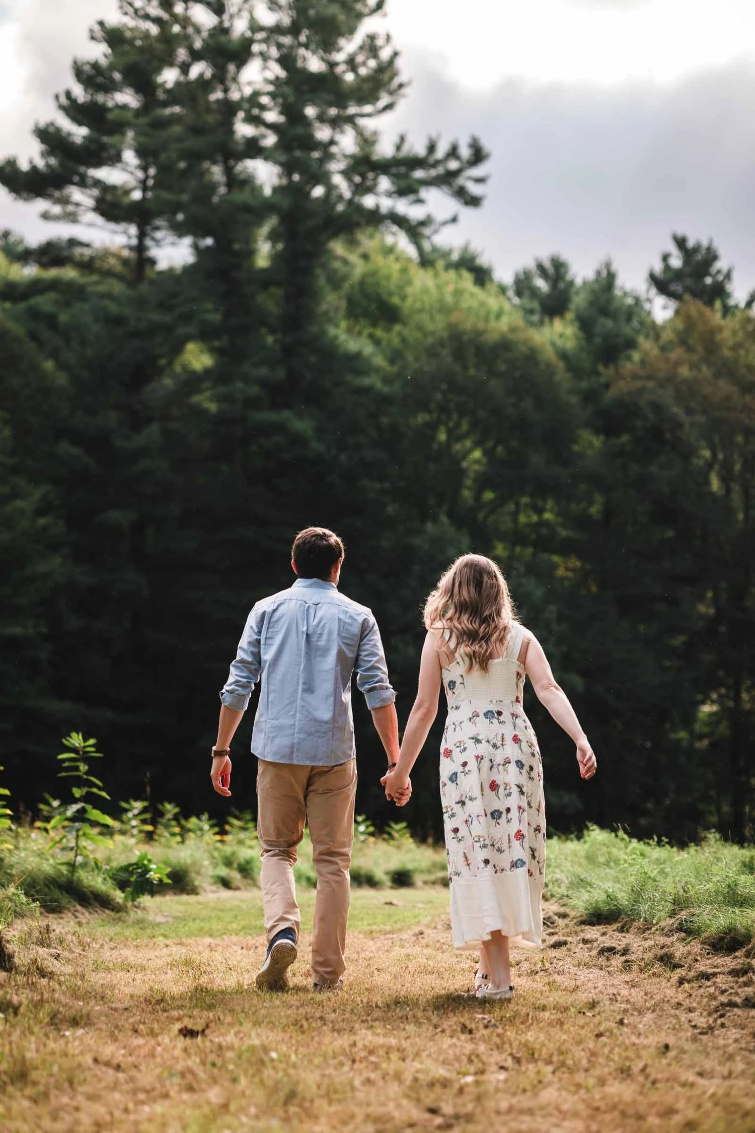 Couple walking hand-in-hand away from camera across a dirt path toward tree line.  Engagement at Borderland State Park, North Easton, MA