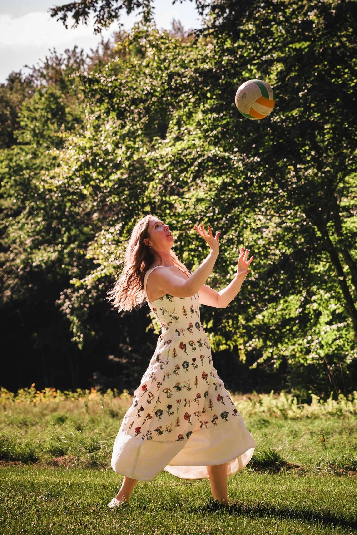 Woman in a floral dress tosses a volleyball in a sunlit garden. Engagement at Borderland State Park, North Easton, MA