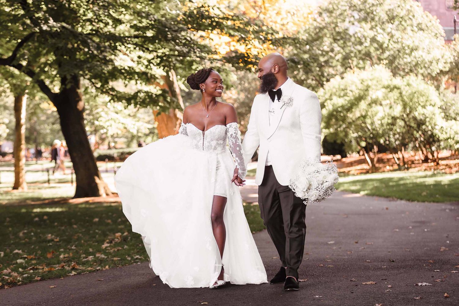 Bride and groom walking hand-in-hand in through the Boston Public Garden, bride in a flowing white gown, groom in a white tuxedo jacket.