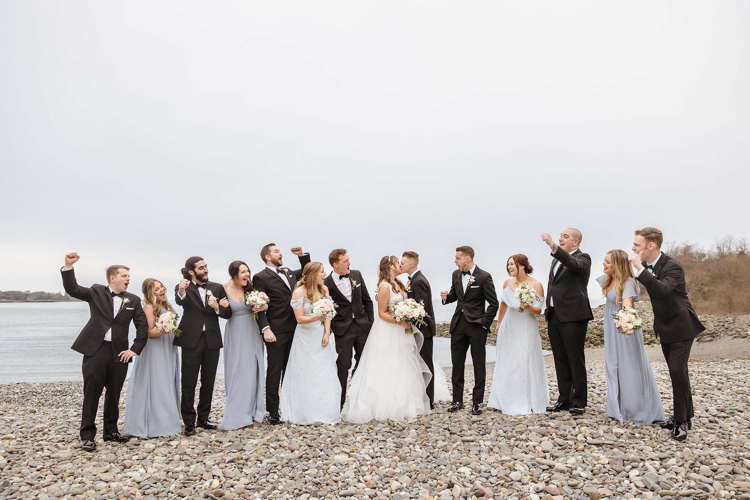The entire wedding party pose for a portrait while cheering the bride and room on with a kiss.