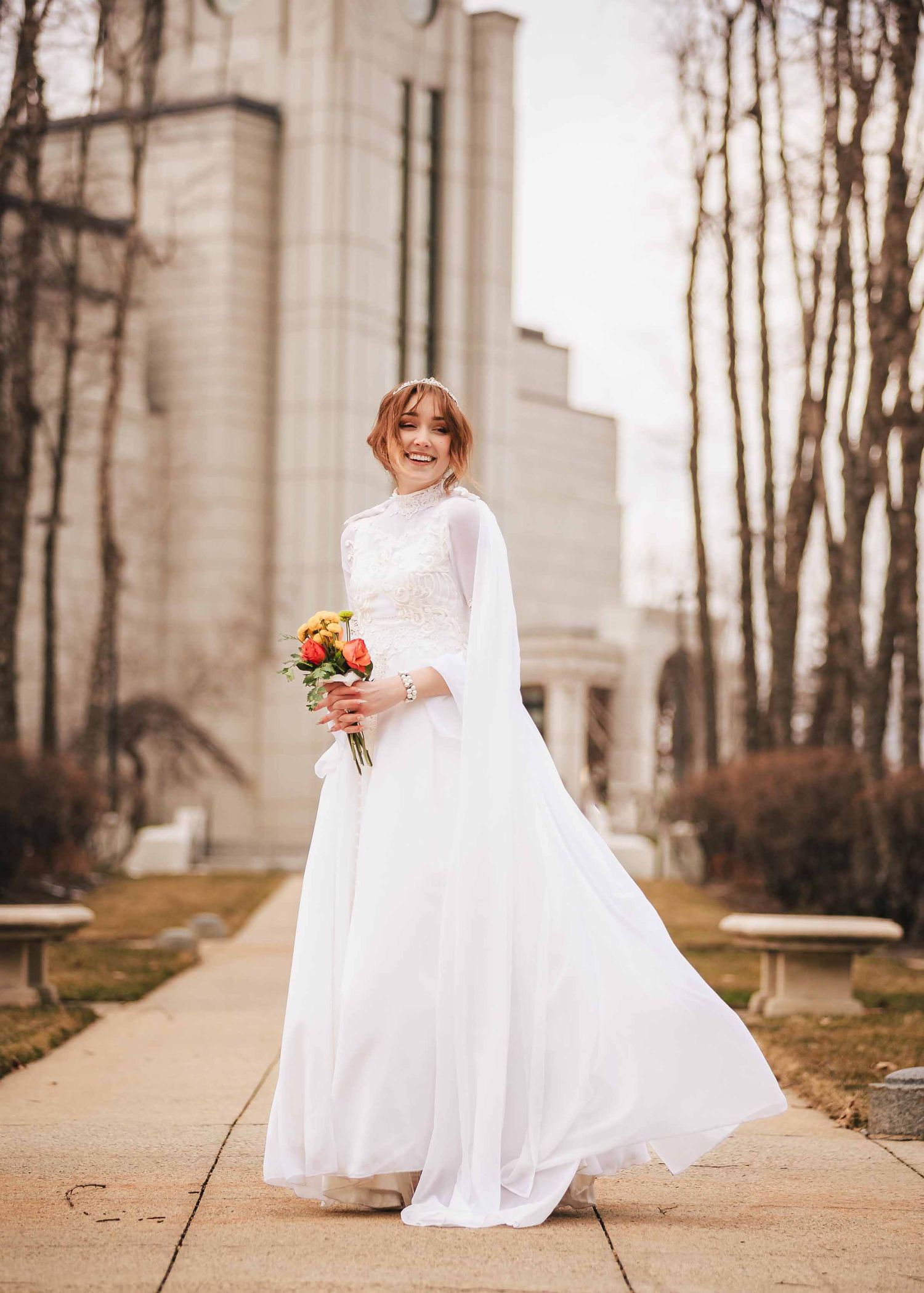 Bride in elopement at Boston Massachusetts Temple holds her bouquet while standing alone in a tree-lined corridor, gazing toward camera