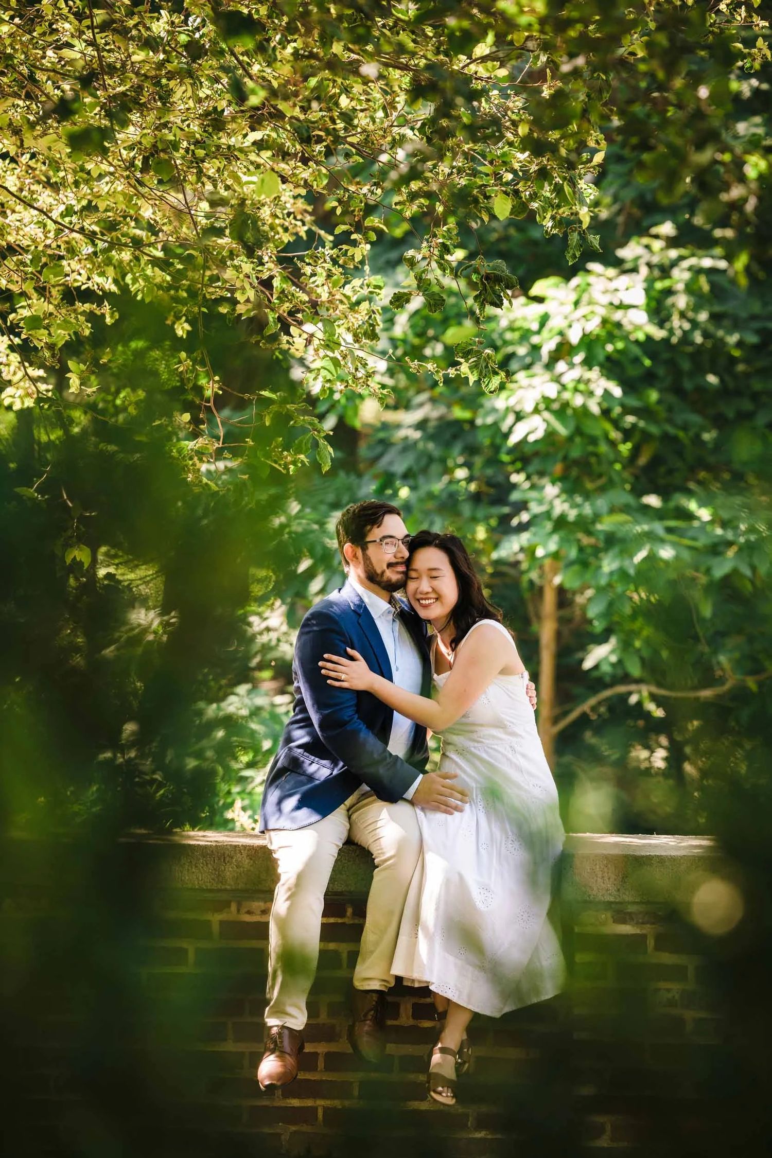 Couple sitting together on a brick bench, embracing and smiling under leafy tree canopy during their engagement portrait session.