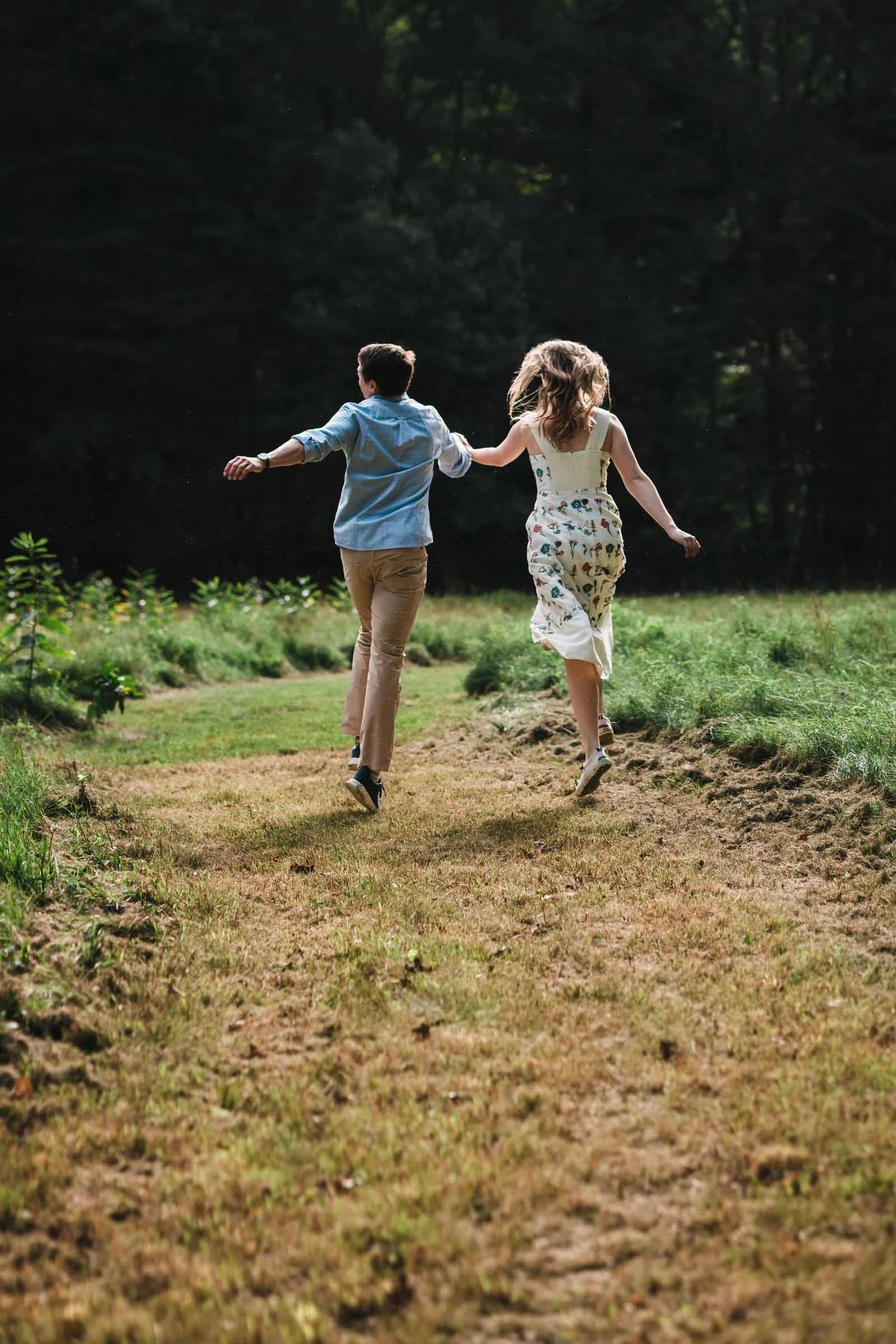 An engaged couple running hand-in-hand down a dirt path through a grassy field. Engagement at Borderland State Park, North Easton, MA