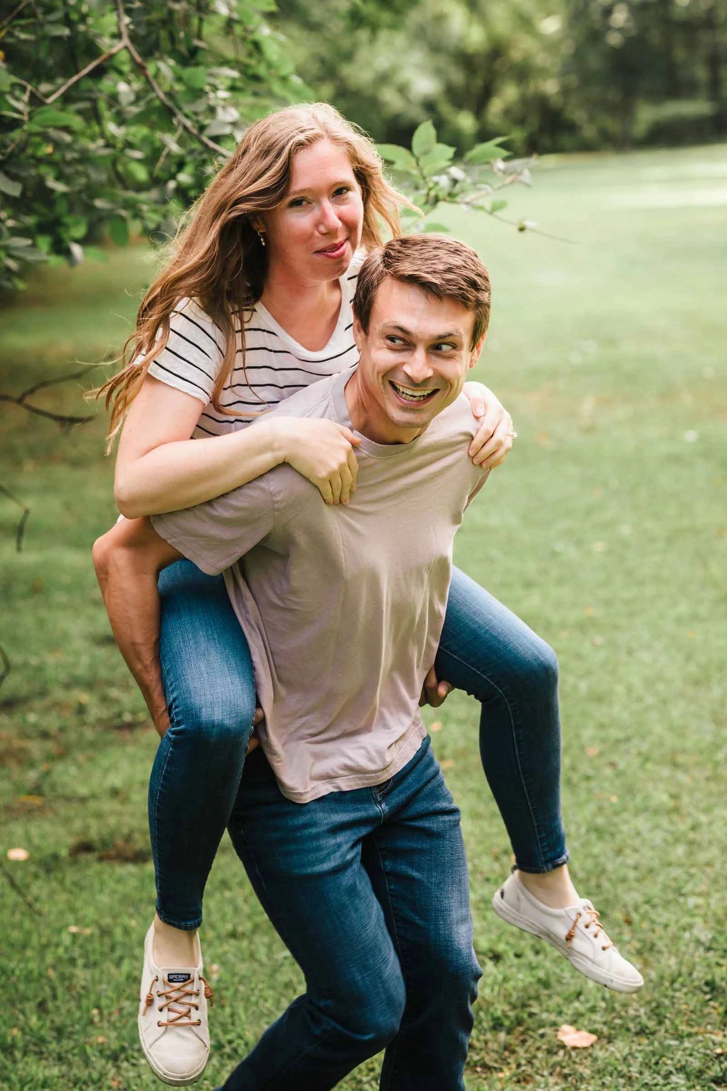 Man giving a woman a piggyback ride while both smile at the camera in a grassy park.  Engagement at Borderland State Park, North Easton, MA
