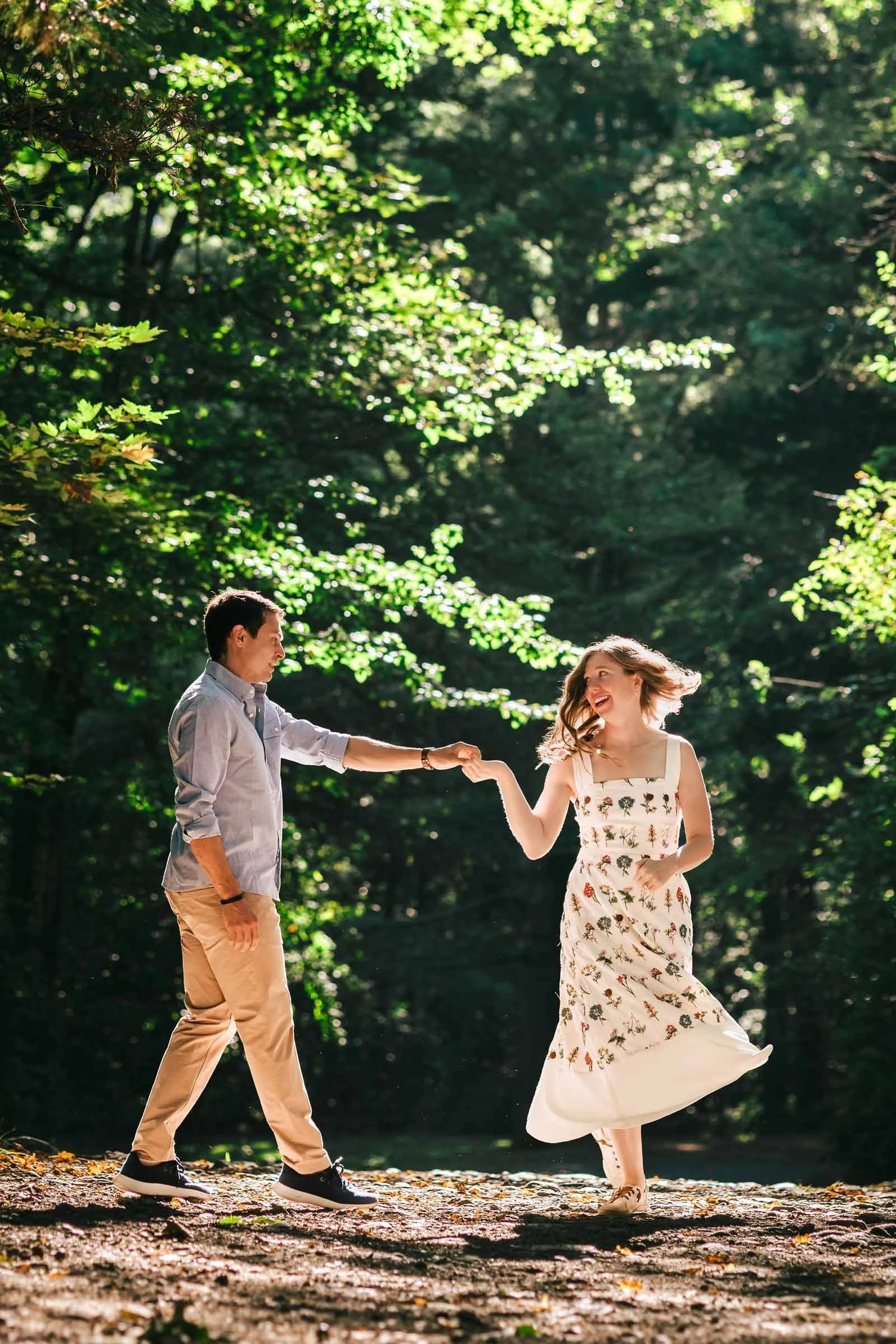 A couple holding hands and dancing together on a forest path dappled with sunlight. Engagement at Borderland State Park, North Easton, MA